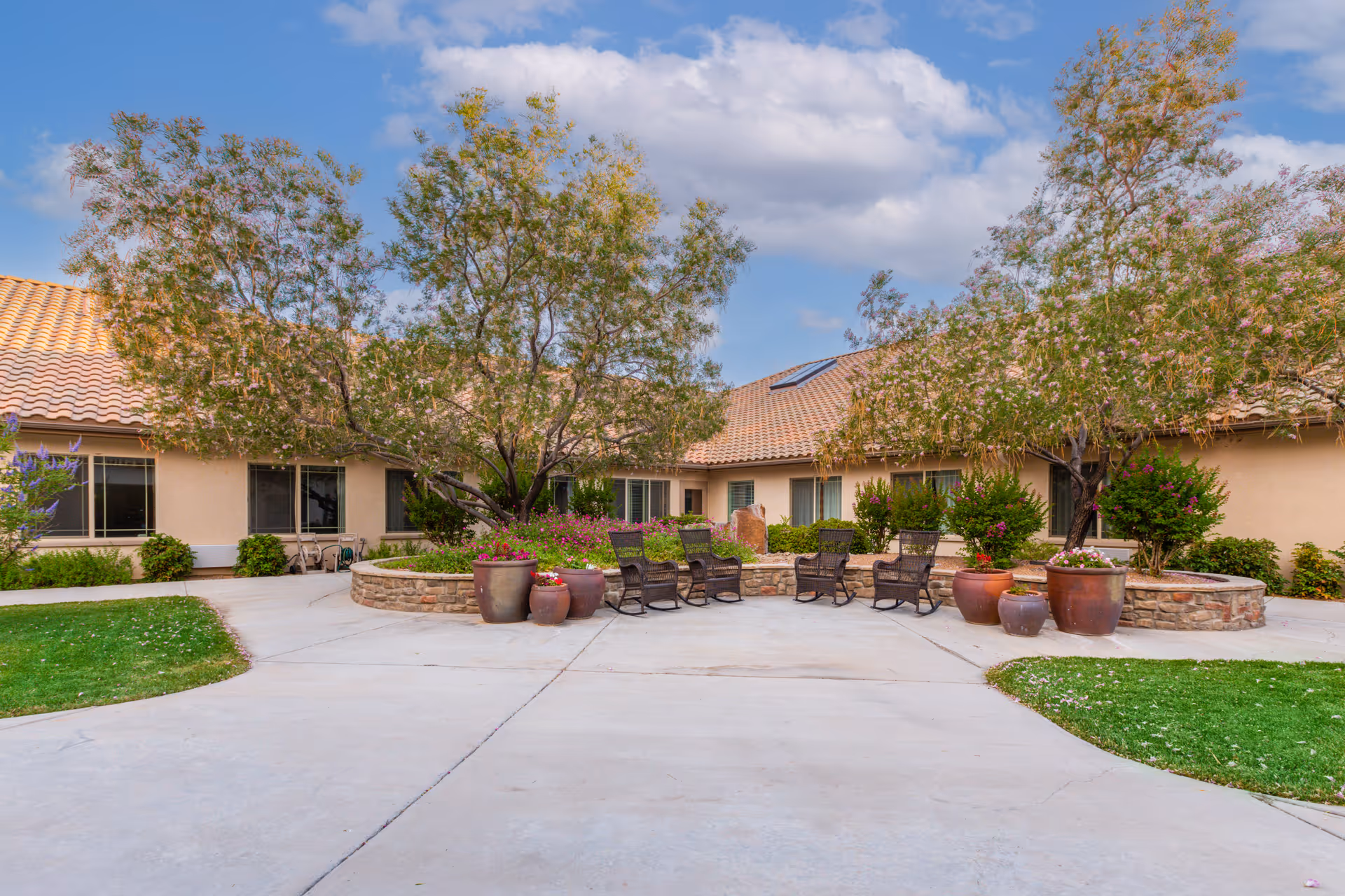Outdoor courtyard area at MorningStar at North Ridge featuring a circular stone planter with trees and flowering plants, surrounded by several wicker rocking chairs. The courtyard is paved with concrete and bordered by green grass. The building with beige walls and a tiled roof is visible in the background under a partly cloudy sky.