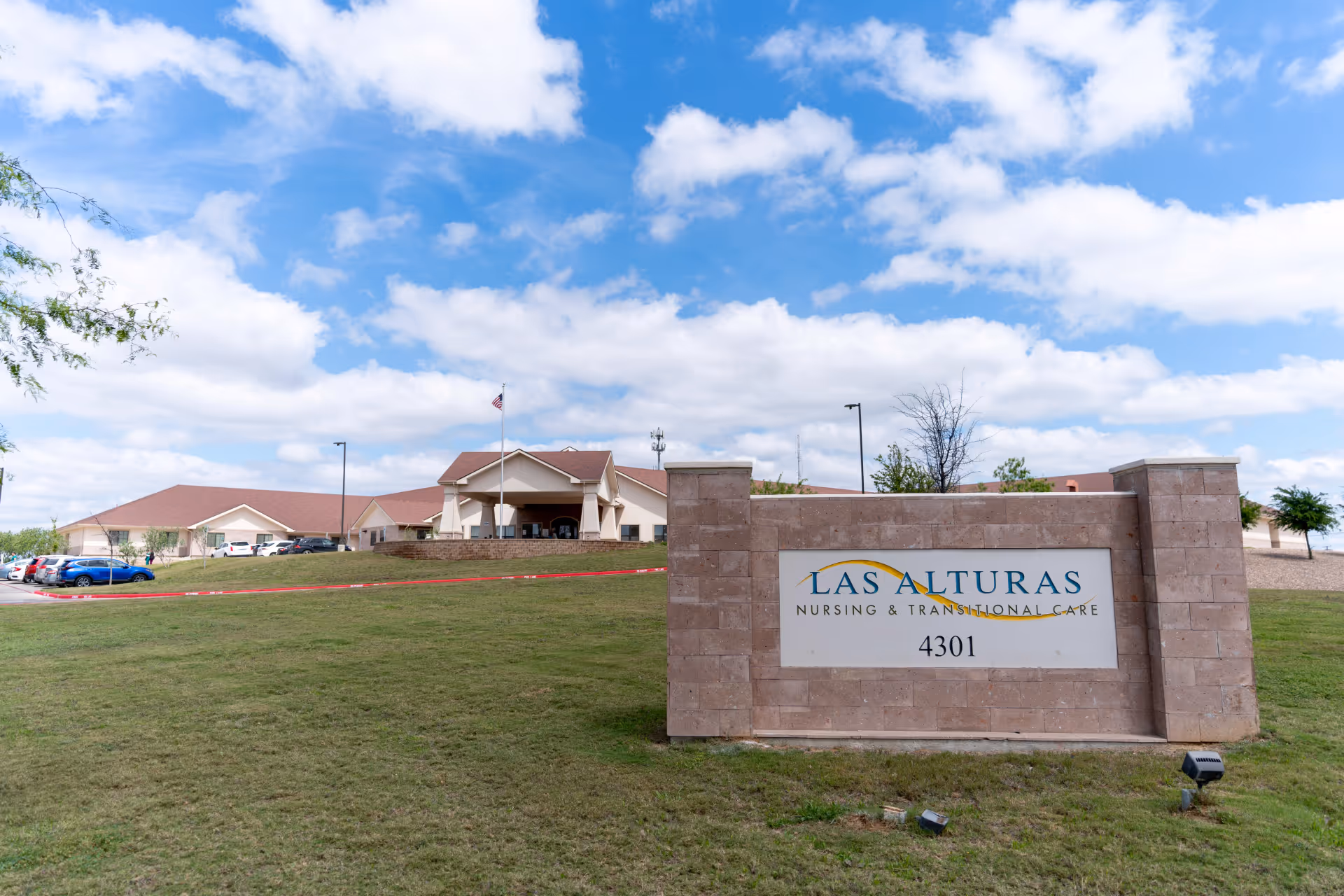 Exterior view of a senior living campus with a stone sign labeled Las Alturas Nursing & Transitional Care in the foreground and a low tan building with a red roof in the background.