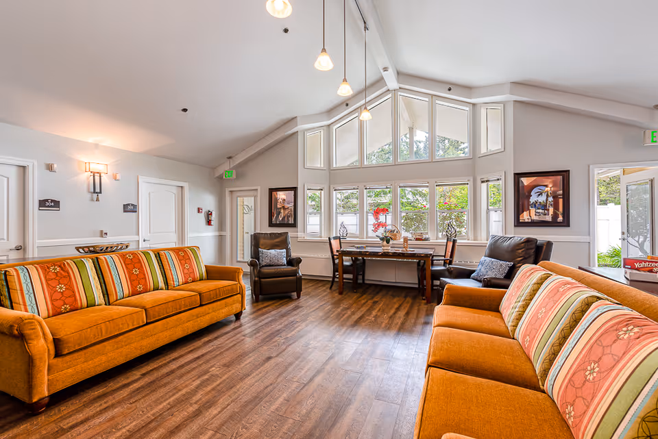 A bright and spacious common area with two orange patterned sofas facing each other, two brown leather armchairs, a wooden table with chairs near large windows, and wood flooring. The room has high ceilings with hanging pendant lights and doors leading to other rooms.