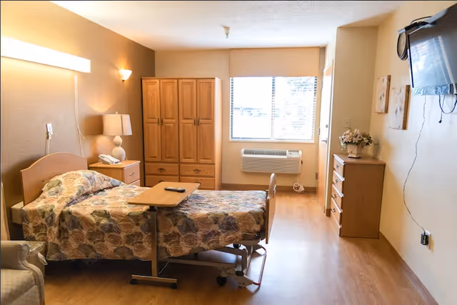 A well-lit retirement community bedroom with a single bed covered in a floral patterned bedspread. The room includes a wooden wardrobe, a bedside table with a lamp and telephone, a small overbed table with a remote control on it, a window with blinds, an air conditioning unit below the window, a wooden dresser with a flower arrangement, and a wall-mounted television with visible cords.