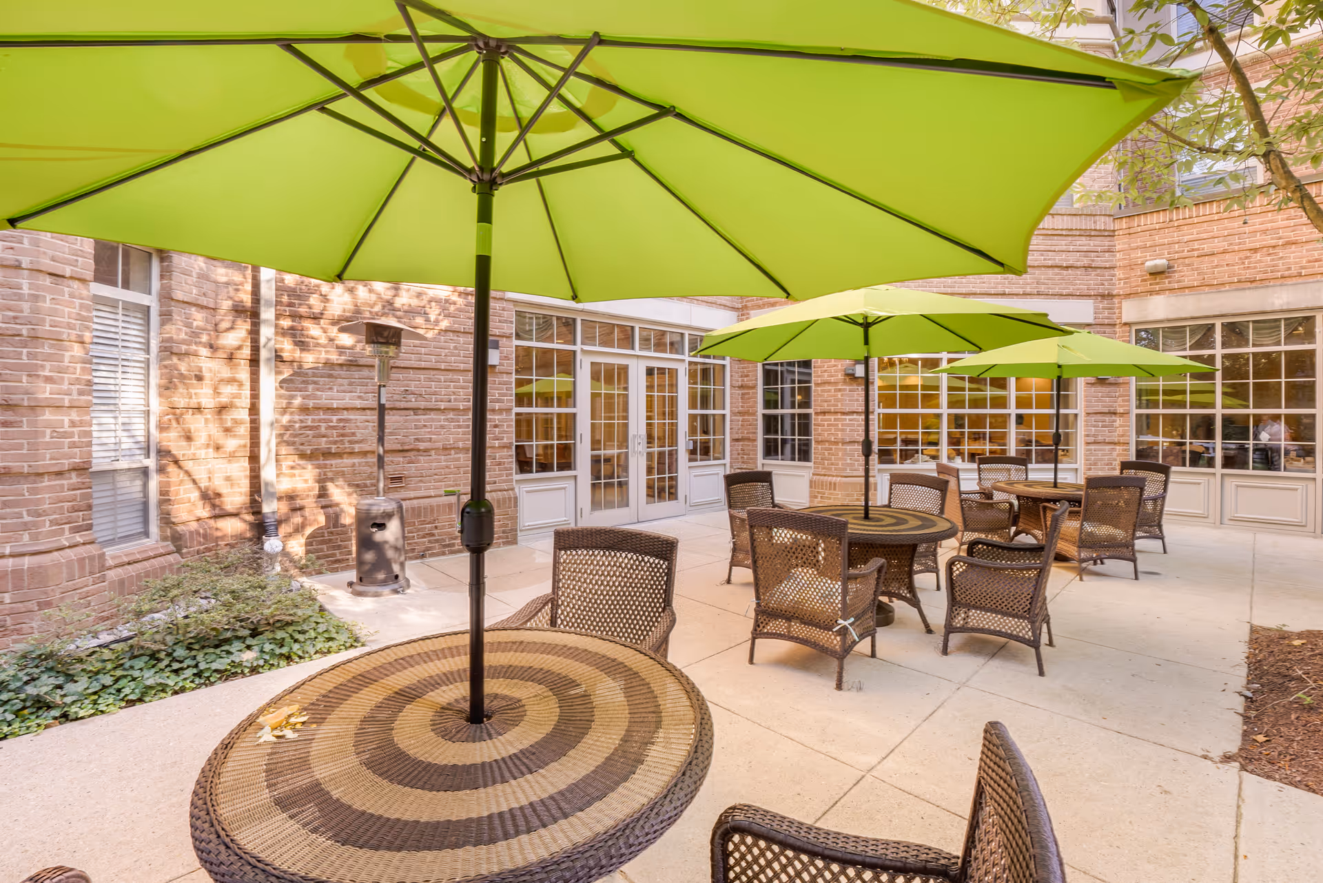 Outdoor patio area with round wicker tables and chairs under large green umbrellas, surrounded by brick walls and windows of the building.