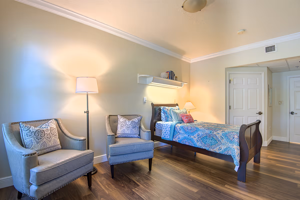 A cozy bedroom in Autumn House featuring a single bed with a colorful blue and turquoise patterned bedspread and multiple pillows. Next to the bed are two gray upholstered armchairs with decorative pillows, and a floor lamp between them. The room has light-colored walls, wooden flooring, a white shelf with books and decor above the bed, and two white doors in the background.