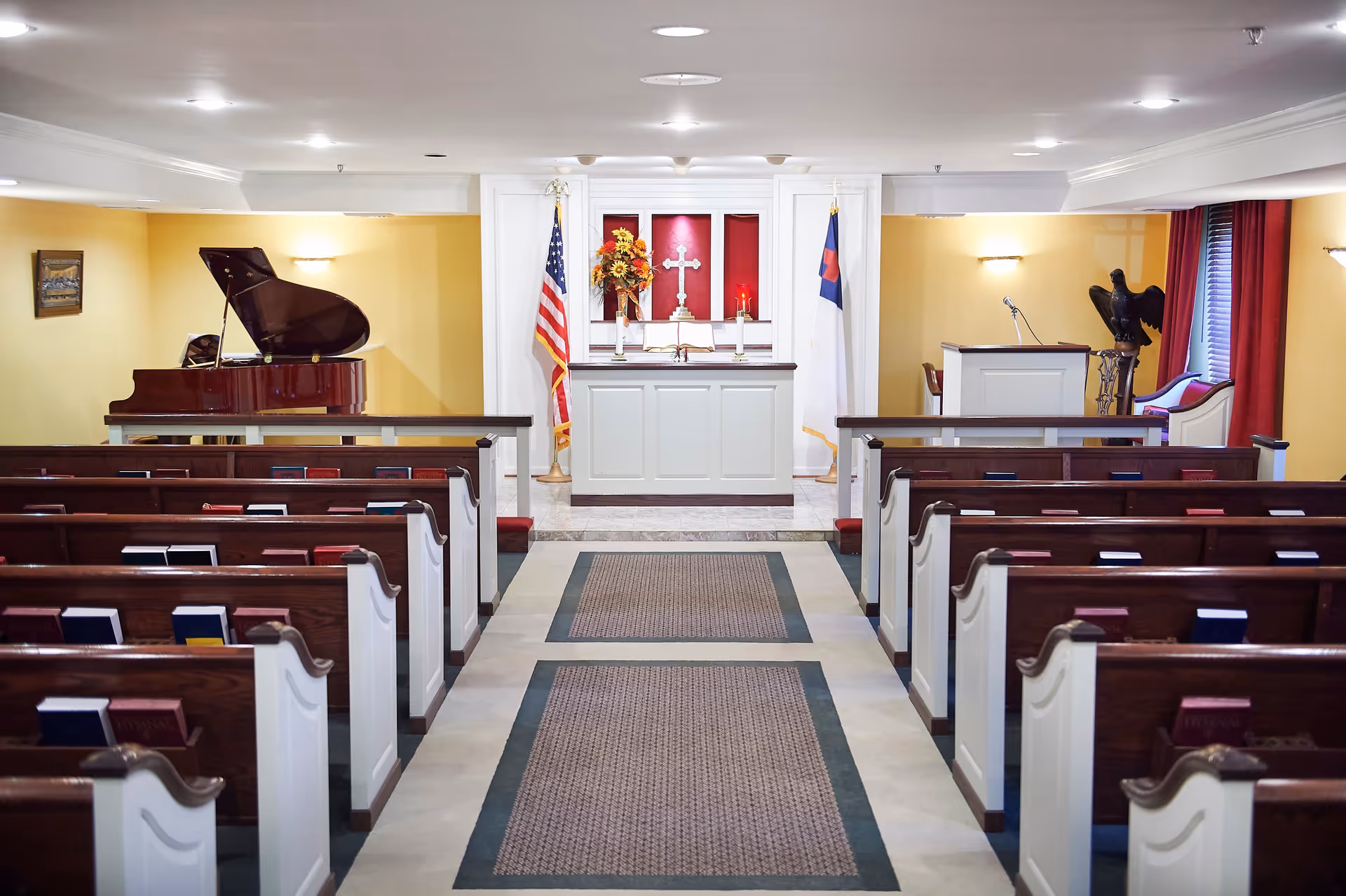 Interior view of a chapel with wooden pews arranged in rows facing an altar. The altar has a cross, candles, an open book, and floral arrangement. Two flags flank the altar, and a grand piano is visible to the left. The walls are painted yellow with white trim, and there is a lectern with a microphone on the right side.