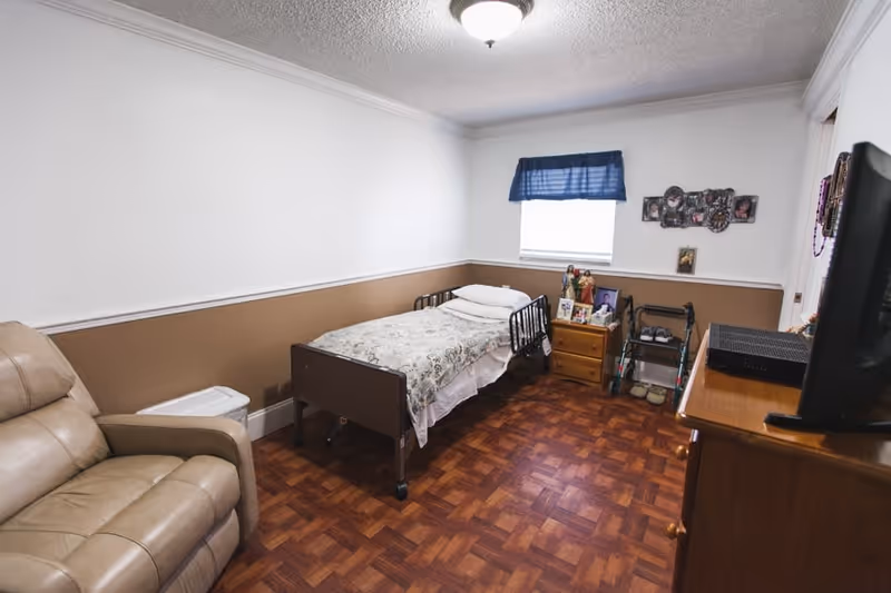 A small senior living bedroom with a single bed featuring a floral bedspread, a beige recliner chair, a wooden dresser with a TV on top, a nightstand with framed photos, a walker, and a window with a blue valance.