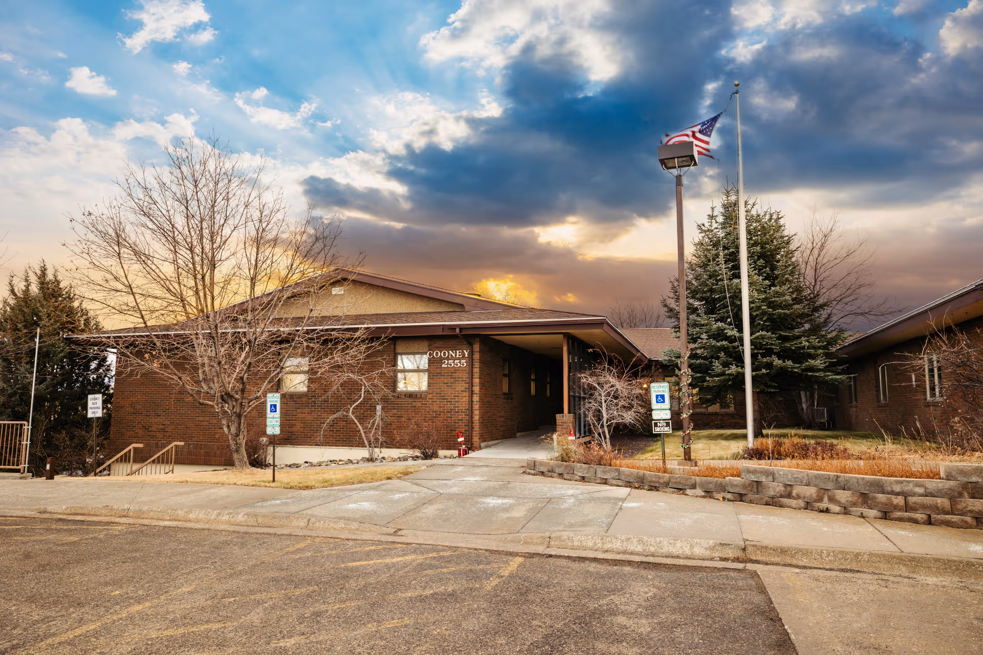 Exterior view of Cooney Healthcare and Rehabilitation building with a brick facade, a flagpole with an American flag, leafless trees, and a cloudy sky at sunset.