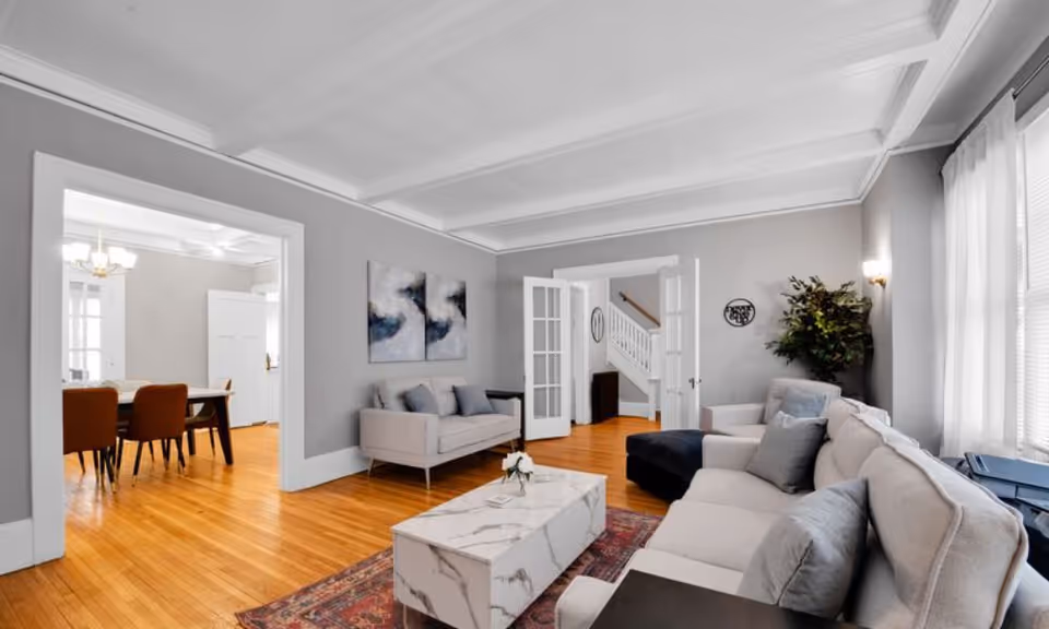 A bright and spacious living room with light gray walls and white ceiling beams. The room features a large beige sectional sofa with gray cushions, a smaller beige loveseat, a marble coffee table on a patterned rug, and a potted plant in the corner. French doors lead to a hallway with a staircase, and an open doorway reveals a dining area with a wooden table and brown chairs.