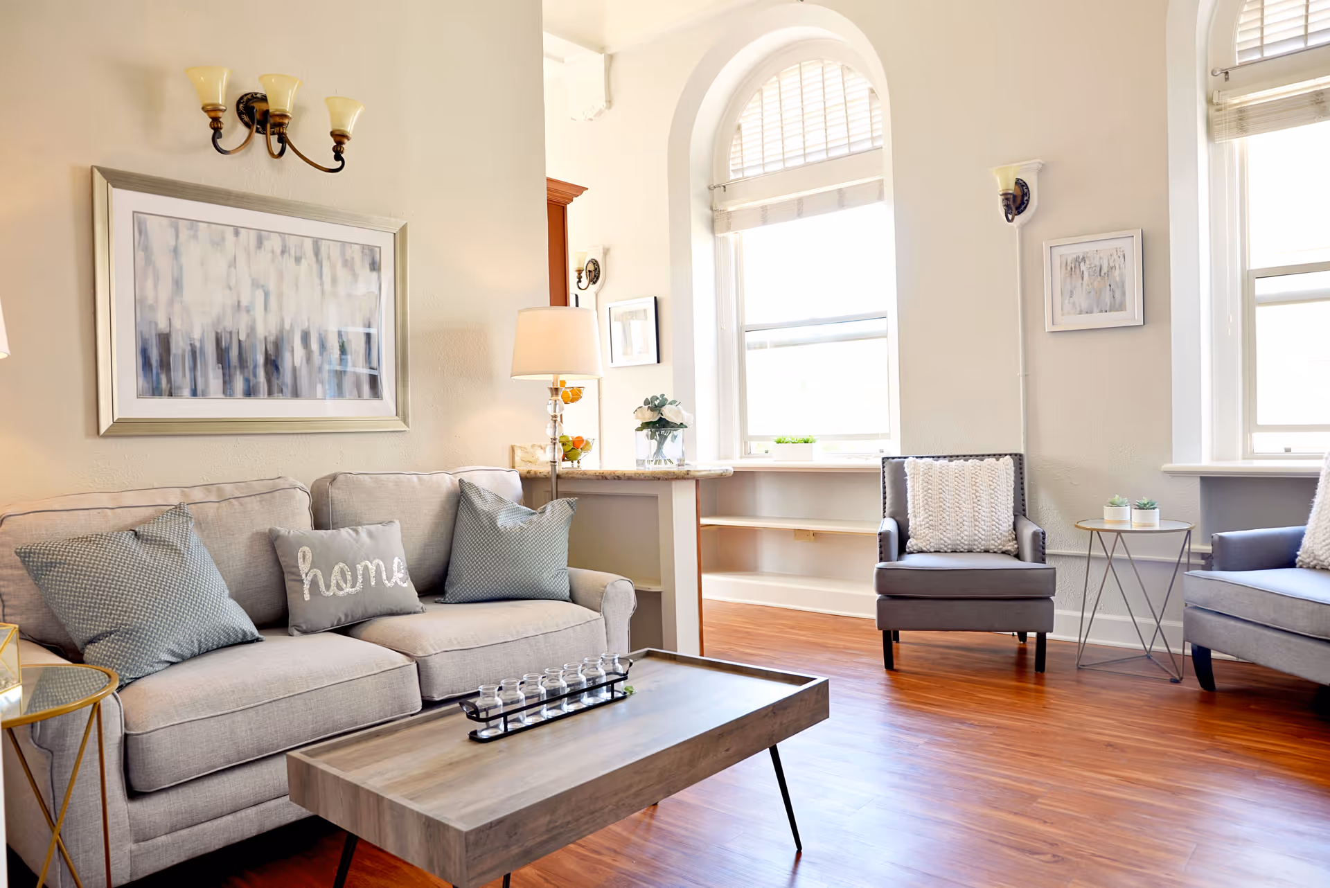 A bright and cozy living room with a light gray sofa adorned with three pillows, one of which says 'home'. There are two gray armchairs with white knitted pillows, a wooden coffee table with a decorative item, and a small round side table with two small plants. The room has large windows with white blinds, wooden flooring, and light-colored walls decorated with framed abstract artwork and wall sconces.