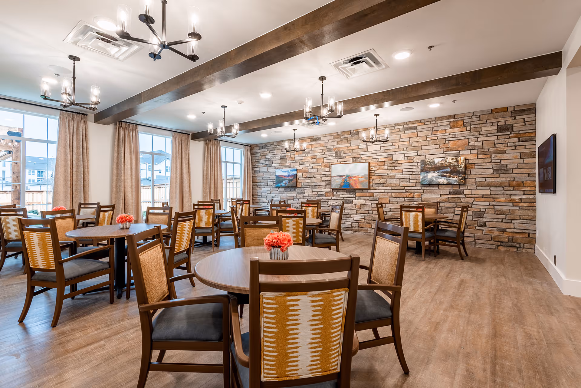 Bright dining room with round wooden tables and chairs, a stone accent wall with artwork, large windows, and chandelier lighting.