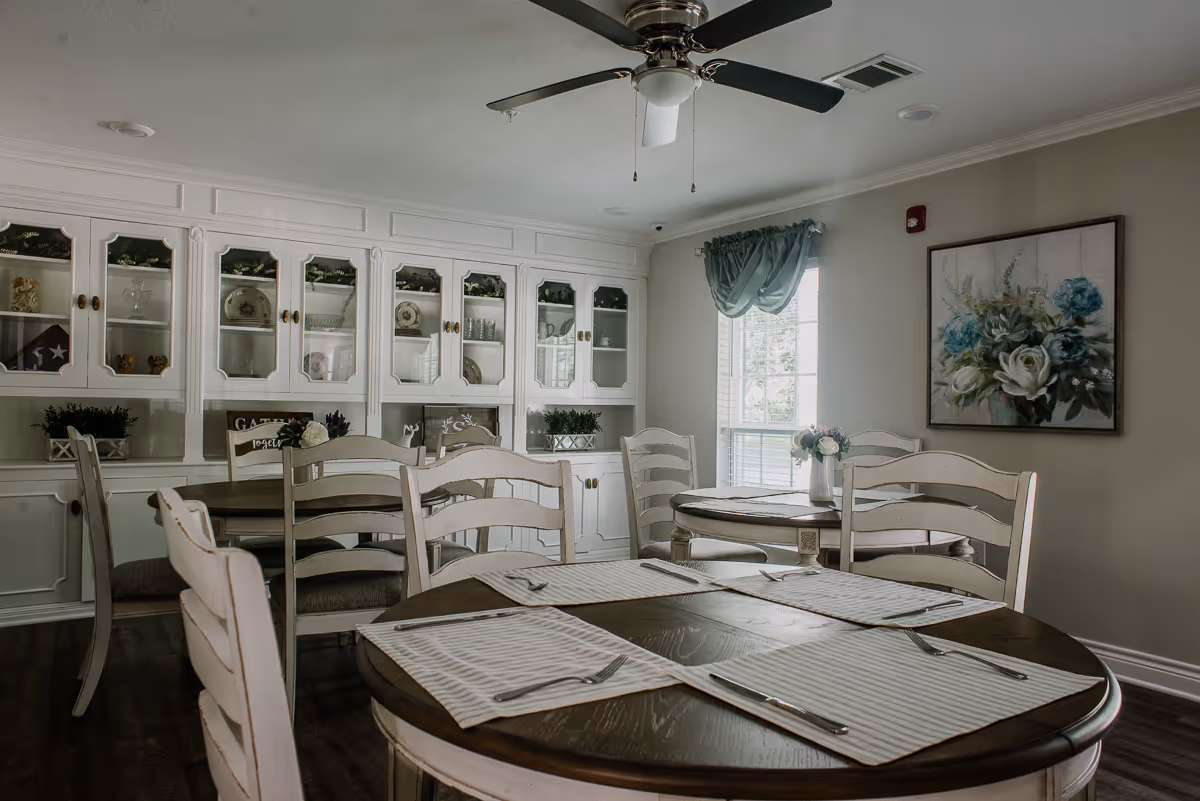 A dining room with round wooden tables set with placemats and silverware. White wooden chairs surround the tables. A large white cabinet with glass doors displaying dishes and decorative items is against the wall. A window with a green curtain and a floral painting are also visible.