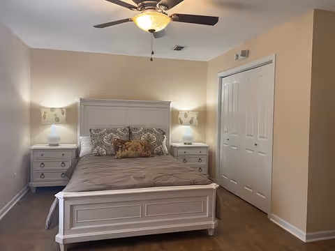 Well-lit bedroom with a white bed frame flanked by matching nightstands and lamps, a ceiling fan, and a double-door closet.