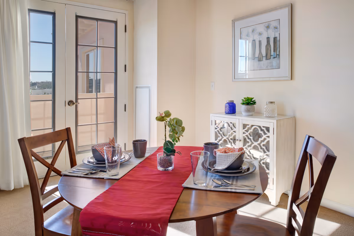 A bright dining area with a round wooden table set for two, featuring plates, bowls, glasses, and napkins. A red table runner decorates the table. Behind the table is a white decorative cabinet with a small plant and jars on top, and a framed artwork hanging on the wall. French doors with glass panes lead to an outdoor balcony, allowing natural light to fill the room.