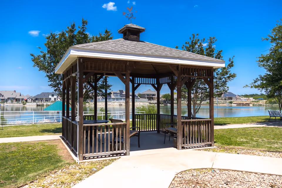 A wooden gazebo with benches inside situated in a grassy area near a lake with houses visible across the water under a clear blue sky.