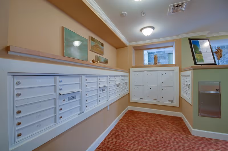 Interior mailroom with rows of white resident mailboxes mounted along peach and green walls above a red-striped carpet.