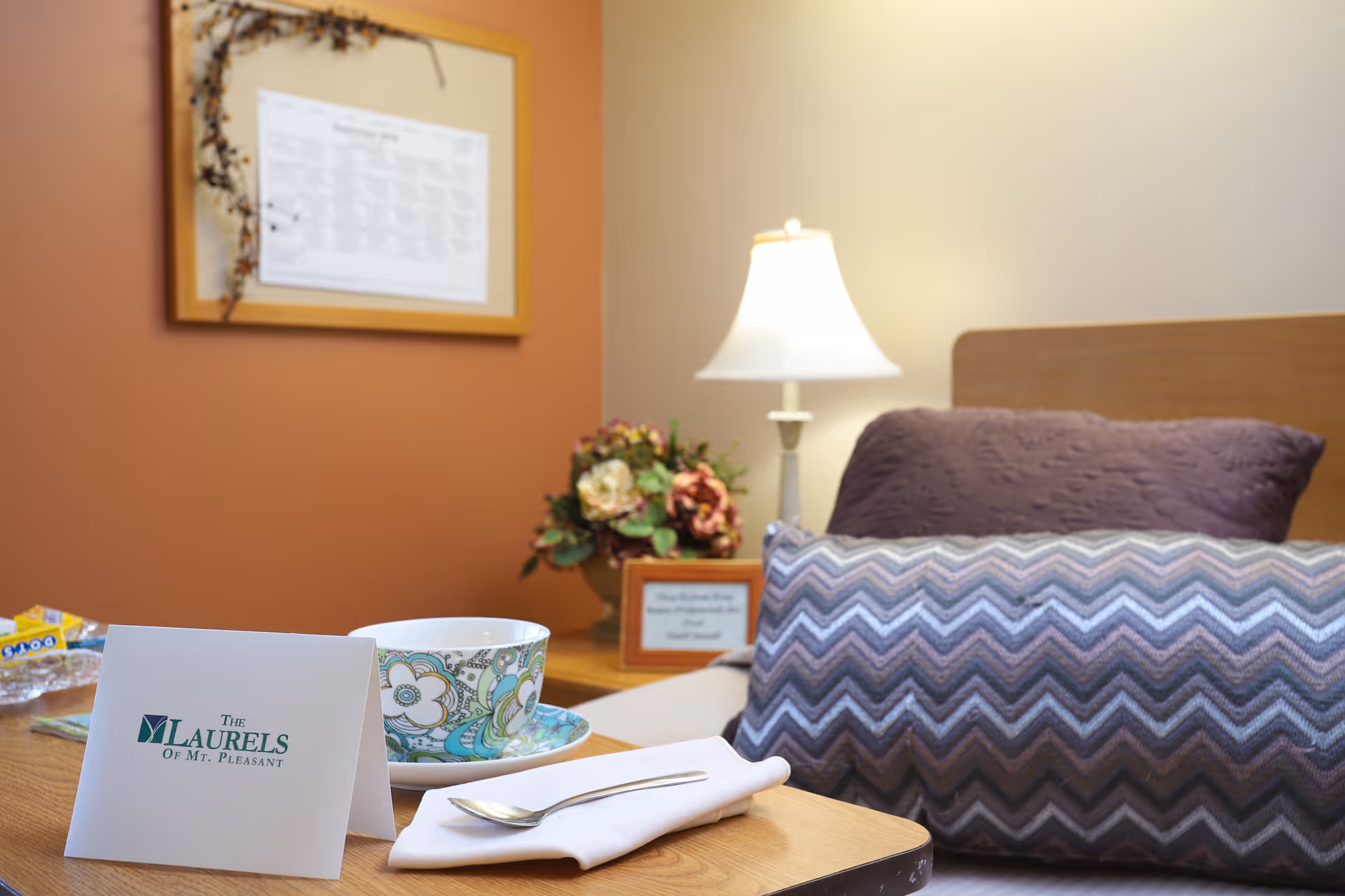 A cozy bedroom corner in a senior living facility with a bed featuring a purple pillow and a chevron-patterned bolster pillow. A wooden bedside table holds a decorative floral arrangement, a lamp with a white shade, and a framed note. In the foreground, a wooden tray holds a patterned teacup and saucer, a folded napkin with a spoon, and a card displaying 'The Laurels of Mt. Pleasant'. The wall behind is painted in warm tones with a framed calendar.
