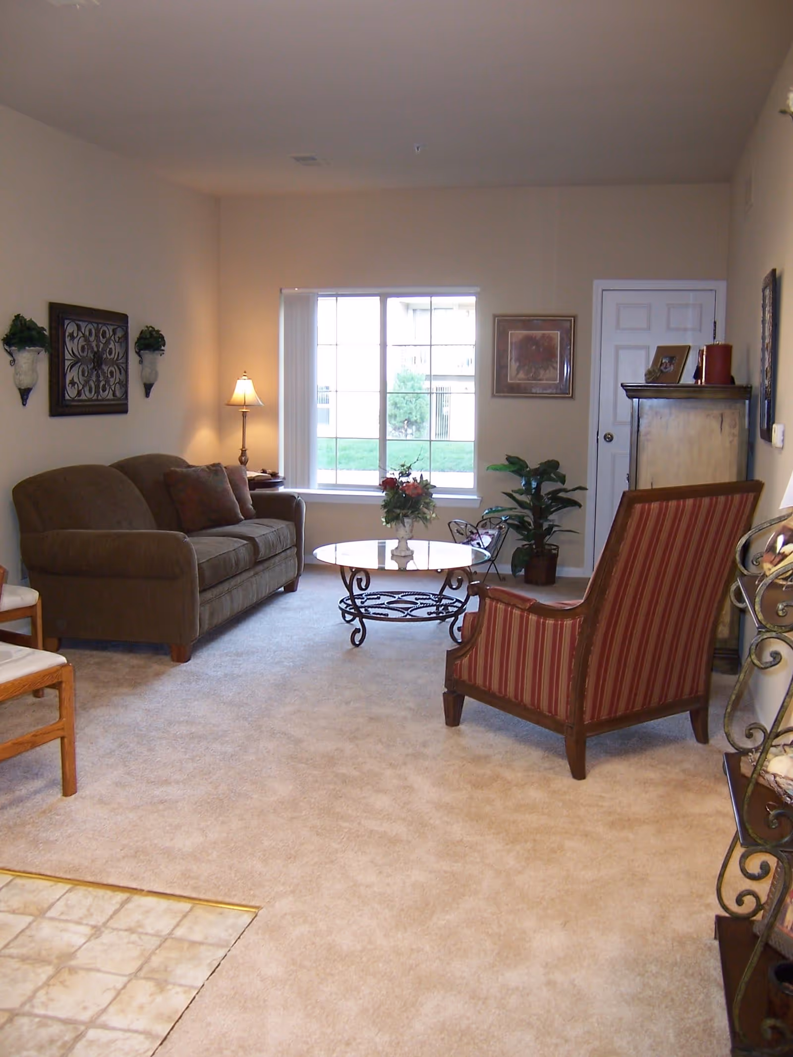A cozy living room with beige carpet, a brown sofa with cushions, a red and brown striped armchair, a round glass coffee table with a floral arrangement, a floor lamp, wall decorations, and a window showing a green lawn outside.