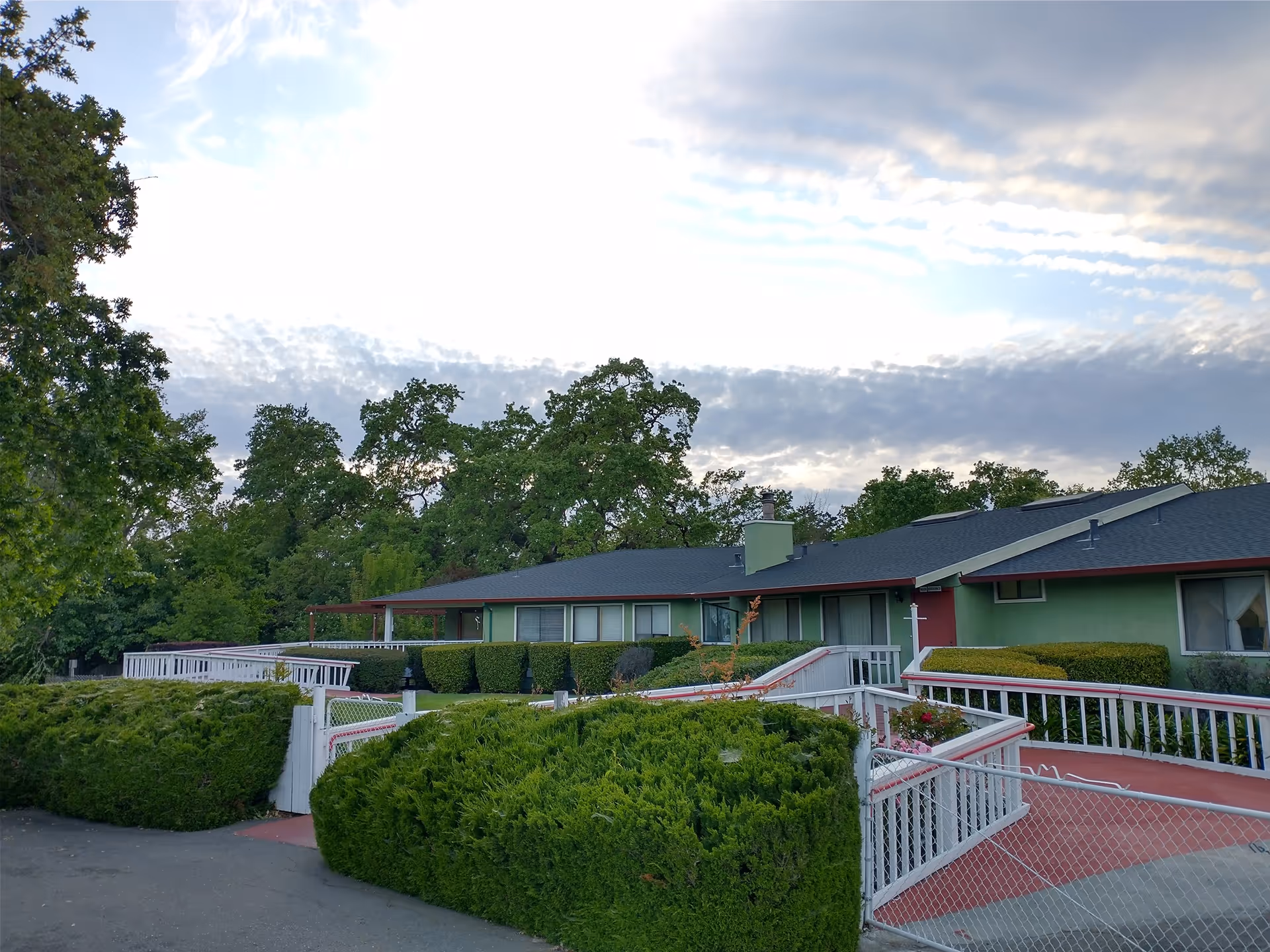 Exterior view of a single-story green building with a dark roof, surrounded by well-maintained bushes and trees under a partly cloudy sky. There are white railings and a gate leading to the entrance.