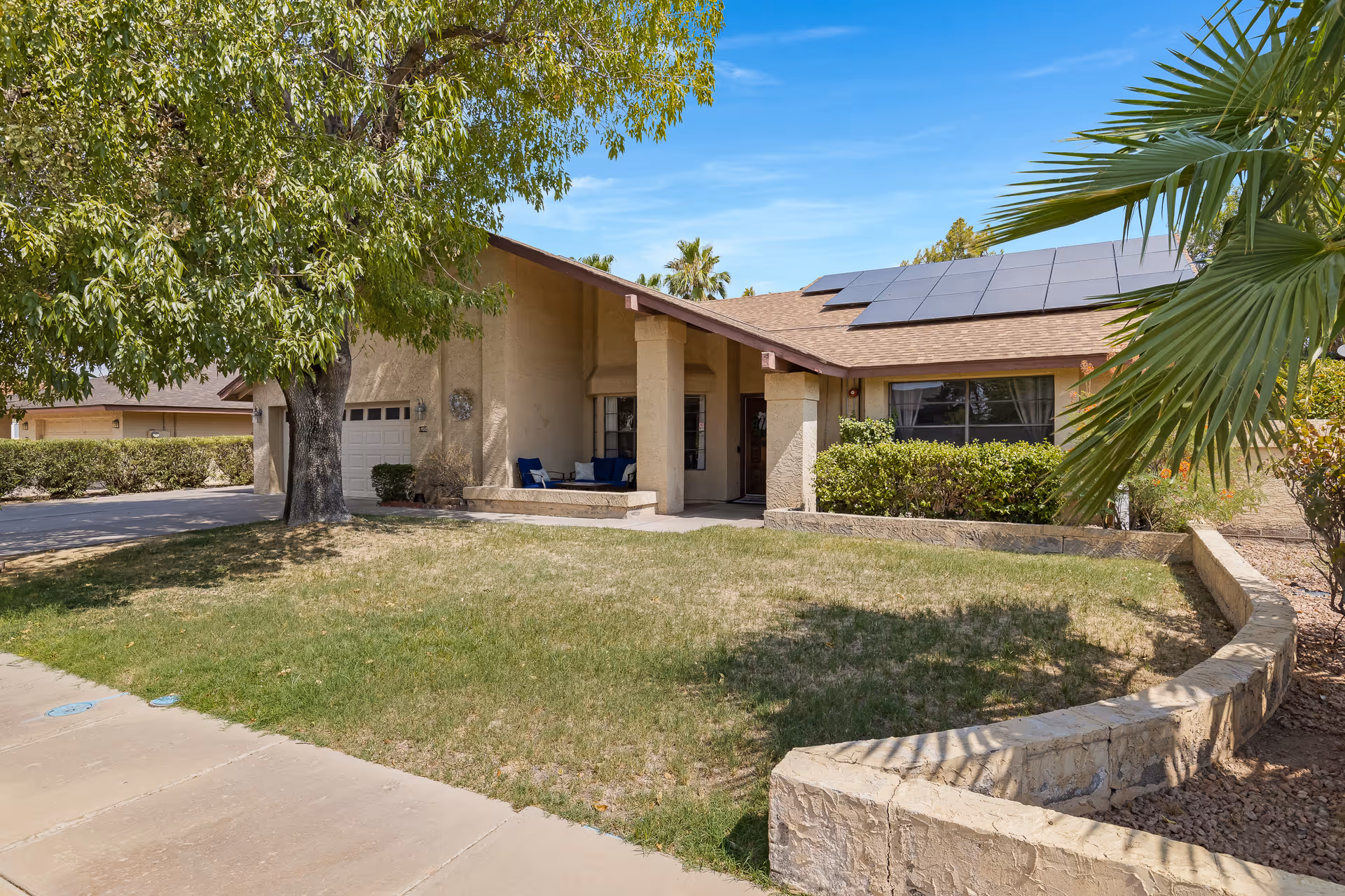 Front exterior view of a single-story house with a covered porch, solar panels on the roof, a large tree on the left, and a palm tree on the right. The yard has a mix of grass and landscaping with a stone border.