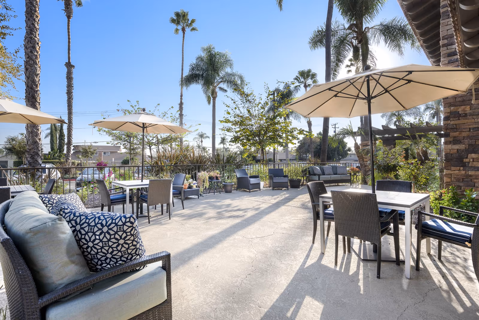 Outdoor patio area with several tables and chairs under large beige umbrellas, surrounded by palm trees and other greenery, with a clear blue sky overhead.