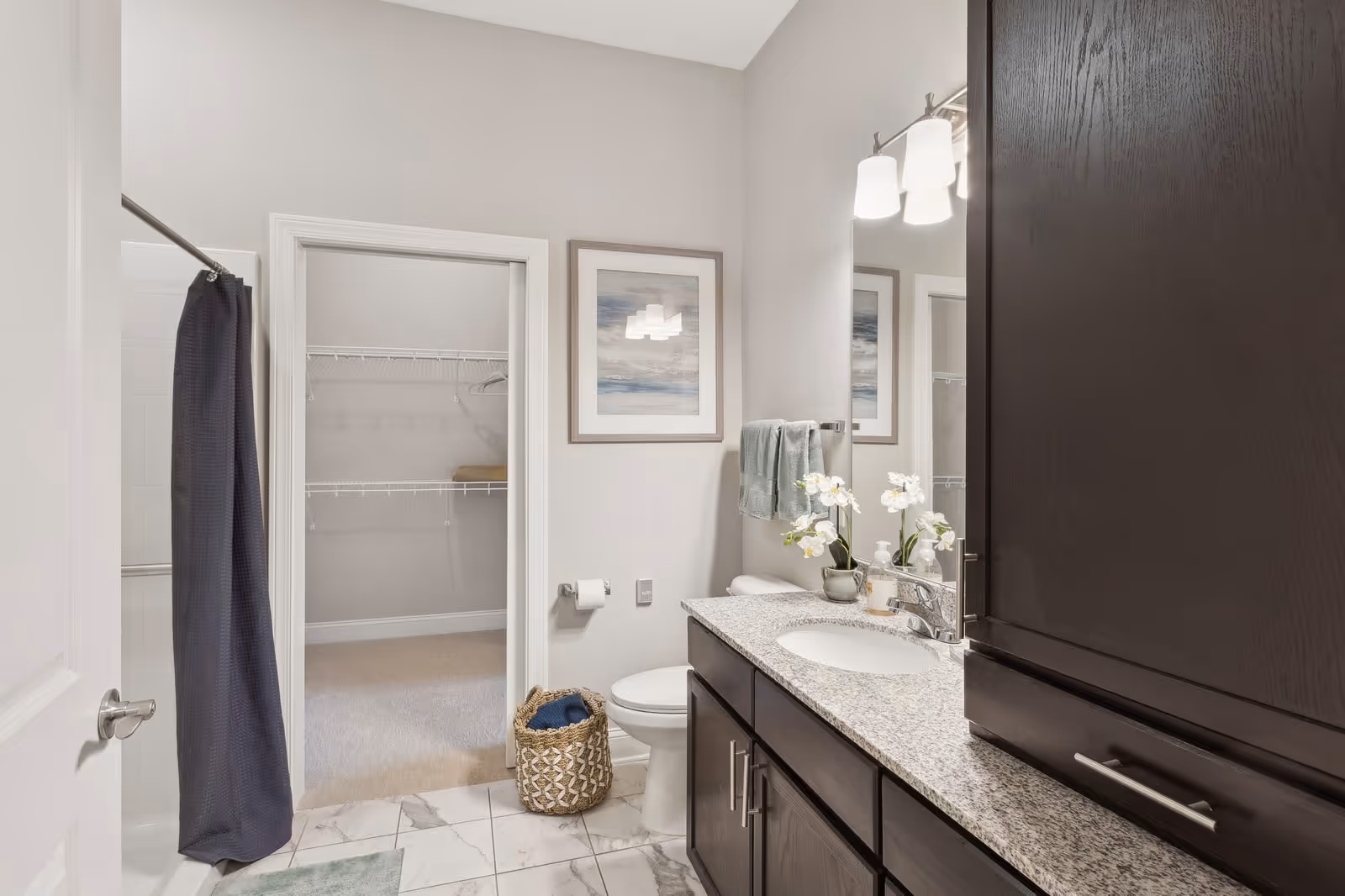 A clean and modern bathroom with a granite countertop vanity, dark wood cabinets, a large mirror, and a sink. There is a toilet next to the vanity with a basket on the floor beside it. A towel rack with two green towels is mounted on the wall above the toilet paper holder. The bathroom has a tiled floor and a shower with a dark curtain on the left side. An open door reveals a walk-in closet with wire shelving. A framed artwork hangs on the wall above the toilet.