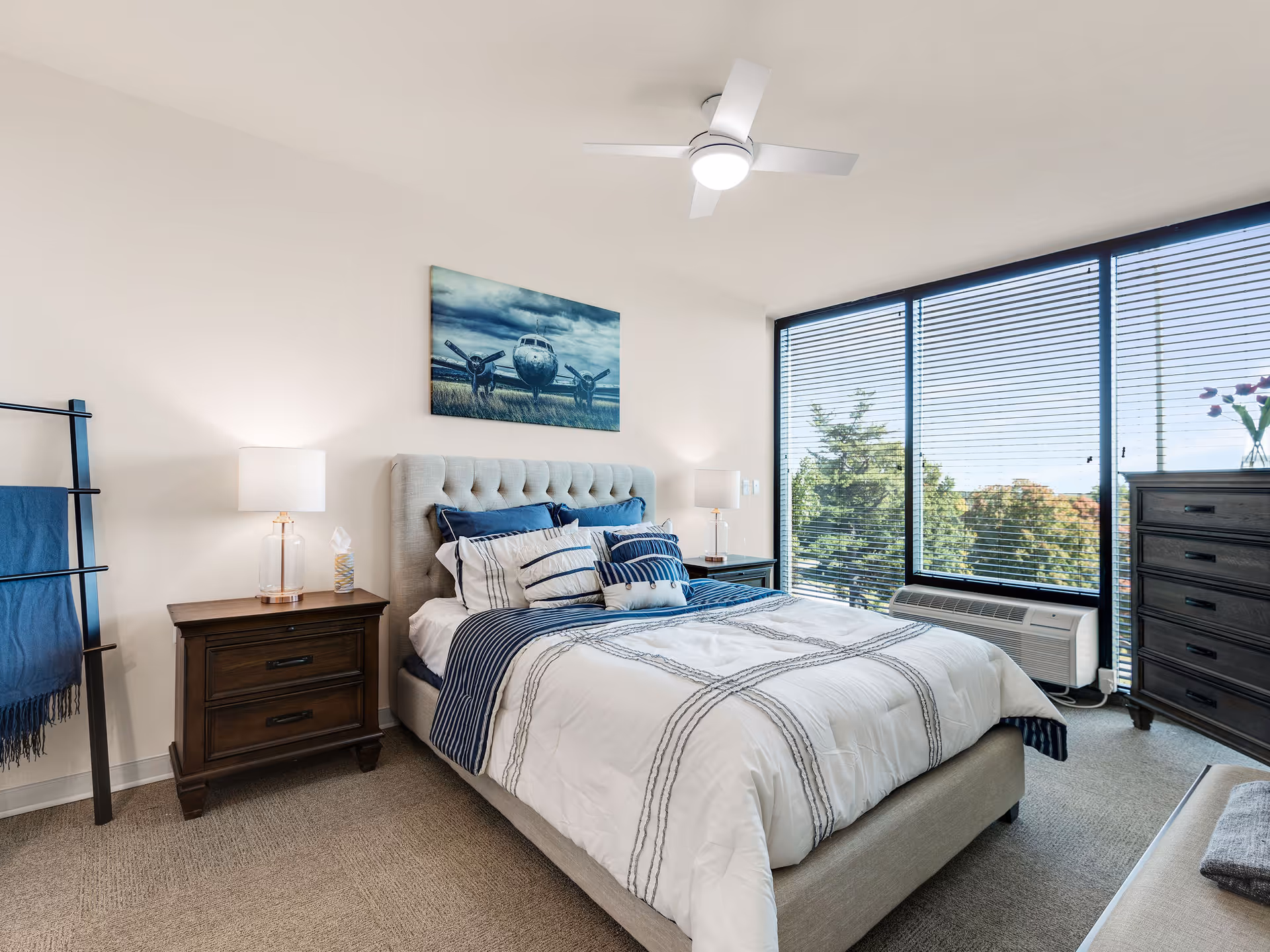 A bright bedroom with a large window covered by blinds showing trees outside. The room features a bed with a tufted headboard, white and blue bedding, two bedside tables with lamps, a dark wooden dresser, a towel rack with a blue towel, and a ceiling fan with a light. A painting of an airplane hangs above the bed.