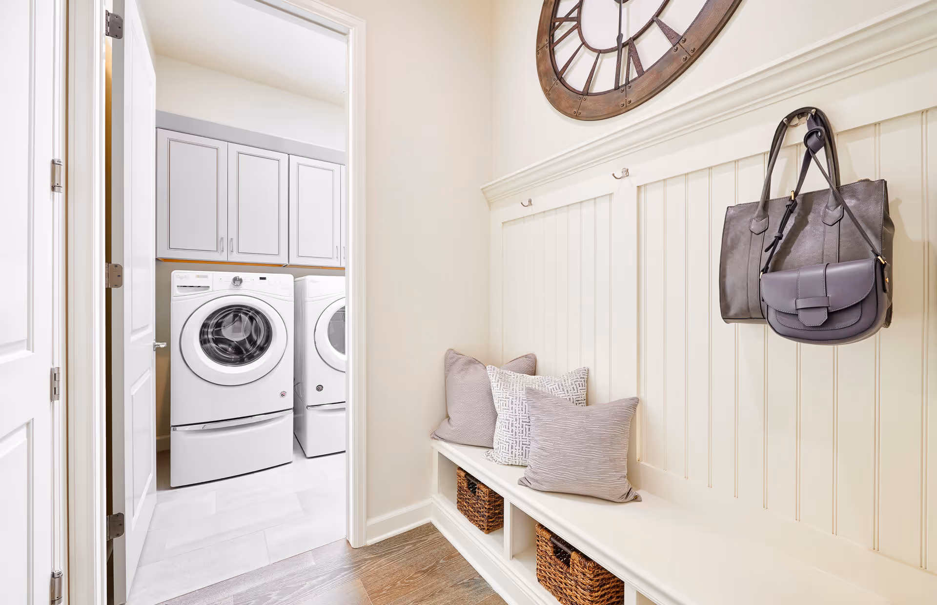 A bright laundry room with a white front-loading washing machine and dryer, visible through an open door. Adjacent to the laundry room is a cozy entryway bench with three decorative pillows and three woven baskets underneath. Above the bench, there are hooks holding two handbags and a large decorative wall clock.