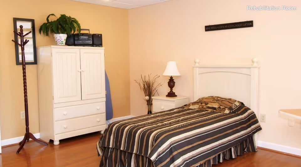 A rehabilitation room with a single bed covered in a striped brown and beige bedspread. Next to the bed is a white nightstand with a lamp and a decorative vase with dried plants. There is a white cabinet with drawers and a potted plant on top, along with a stereo system. A wooden coat rack stands beside the cabinet. The walls are painted beige, and a small sign above the bed reads 'LIVE WELL • LOVE MUCH • LAUGH OFTEN'.