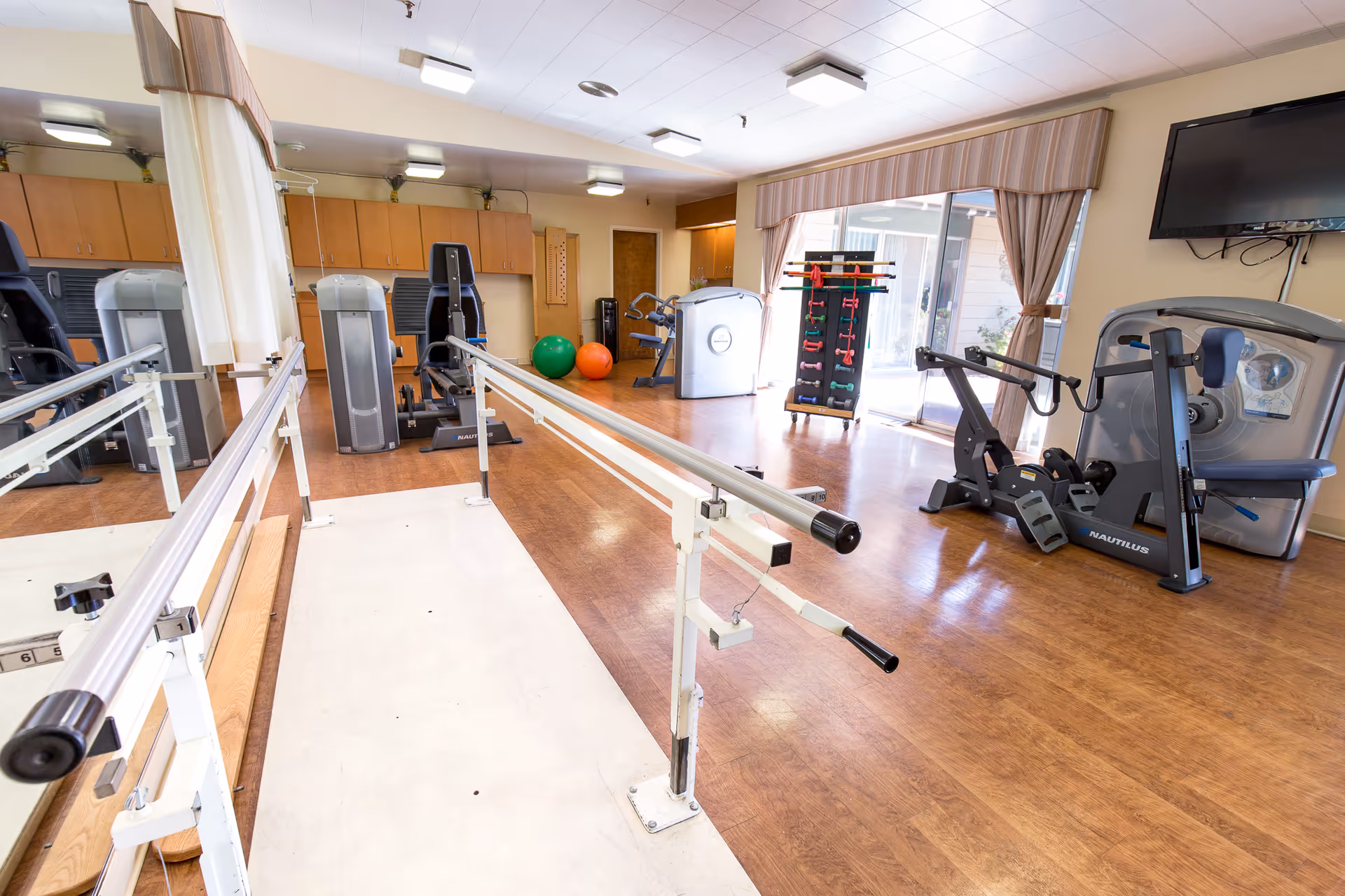 A rehabilitation exercise room with parallel bars for walking practice, various exercise machines, colorful exercise balls, a rack with hand weights, a large mirror on one wall, and a wall-mounted TV. The room has wooden flooring and large windows with curtains allowing natural light to enter.