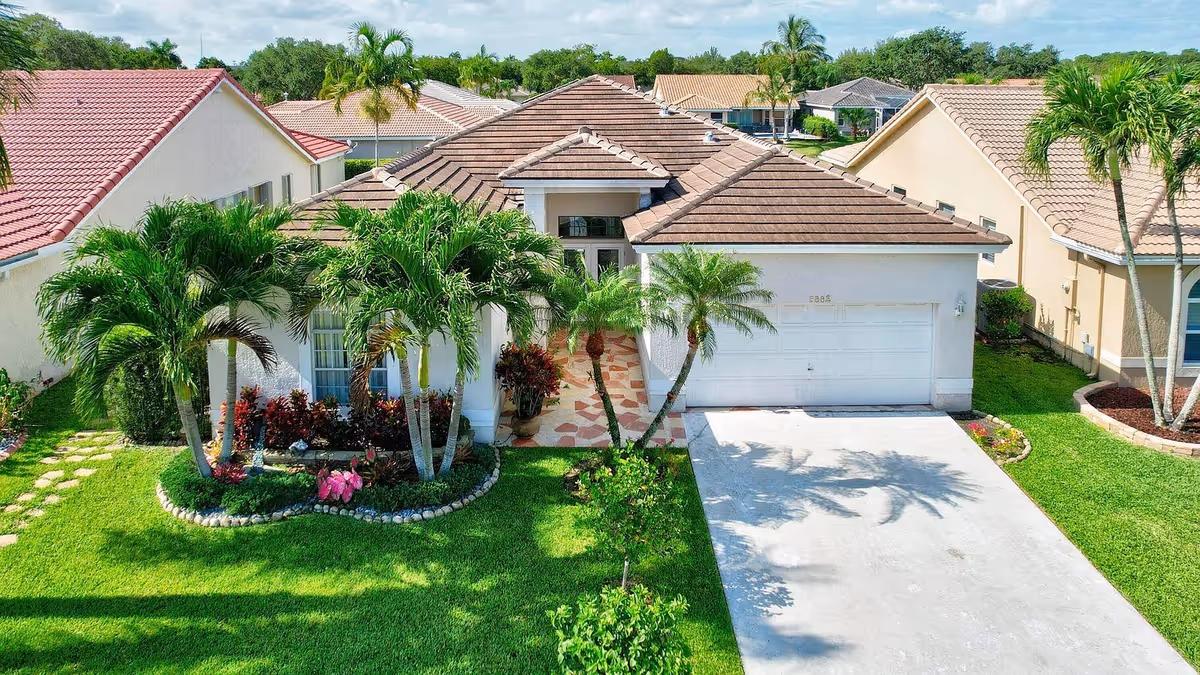 Front exterior of a single-story house with a tiled roof, garage, driveway and palm trees on a manicured lawn.