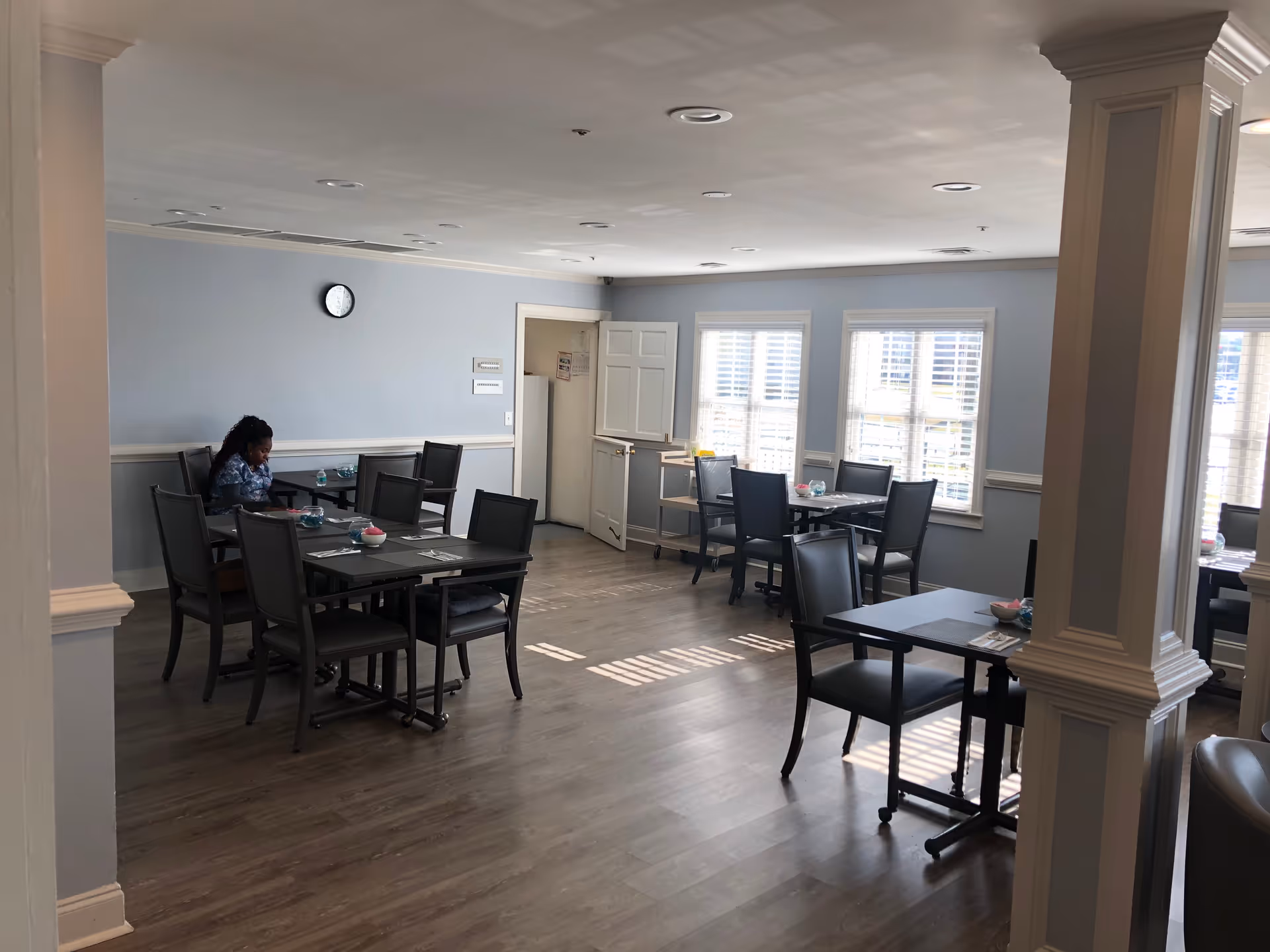 A senior living facility dining room with several tables and chairs arranged neatly. A woman is seated at one of the tables on the left side of the room. The room has light blue walls, large windows with white blinds allowing natural light to enter, and a clock on the wall. The floor is wooden, and there are decorative columns in the room.