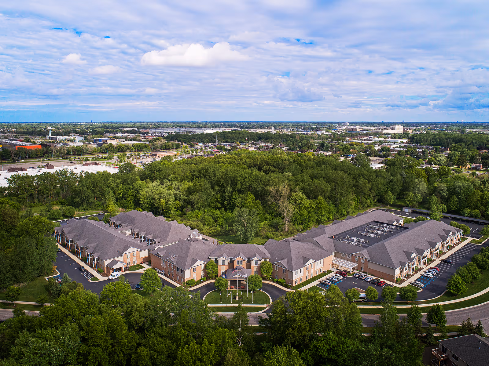 Aerial view of the American House Park Place senior living facility surrounded by trees and greenery, with parking lots and a road visible around the building under a partly cloudy sky.