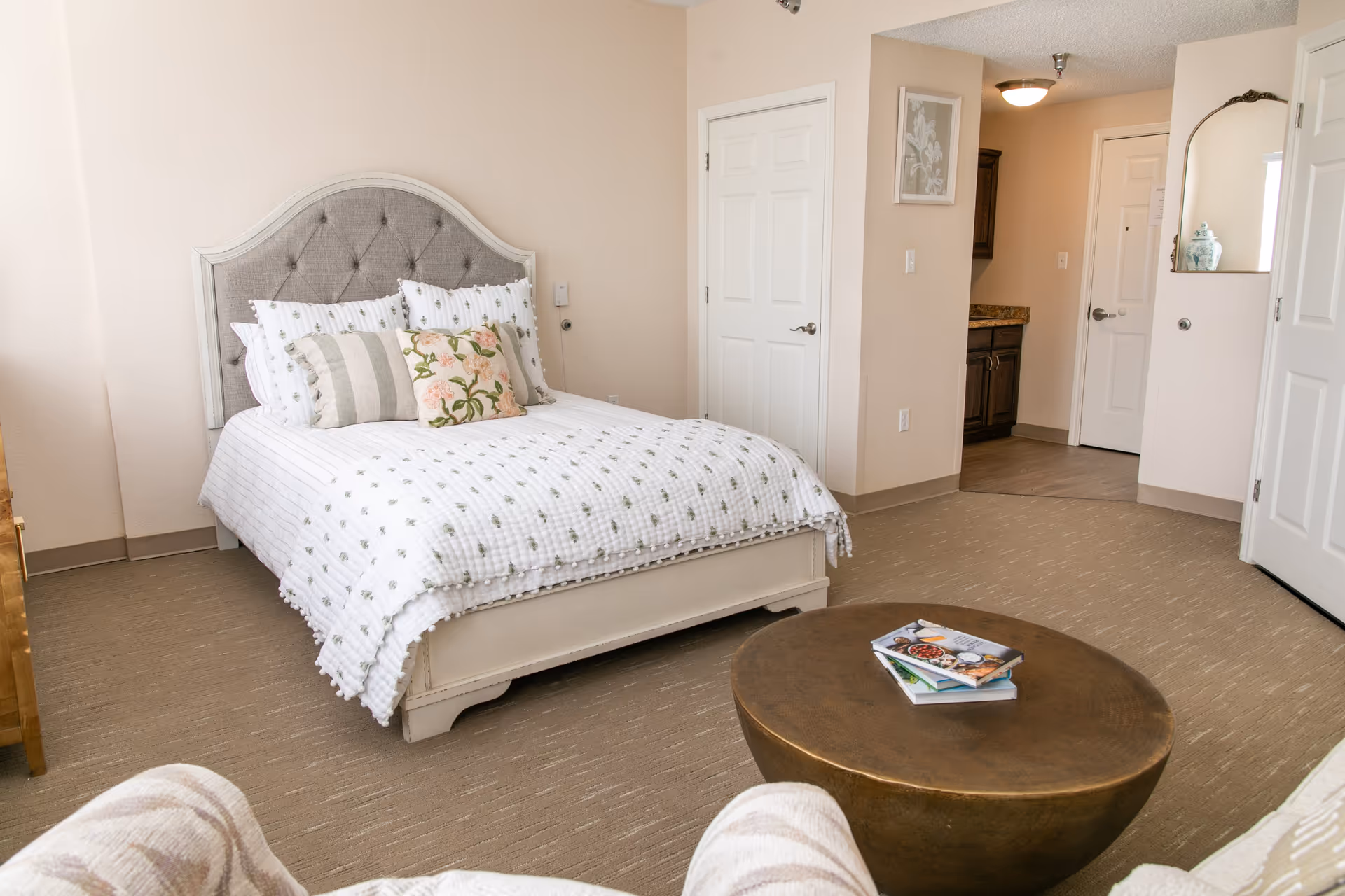 A cozy senior living bedroom with a white bed featuring a tufted gray headboard and multiple pillows. The room has beige walls and carpet, a round wooden coffee table with books on it, and a glimpse of a small kitchenette area in the background. There is a mirror on the wall and several closed doors.