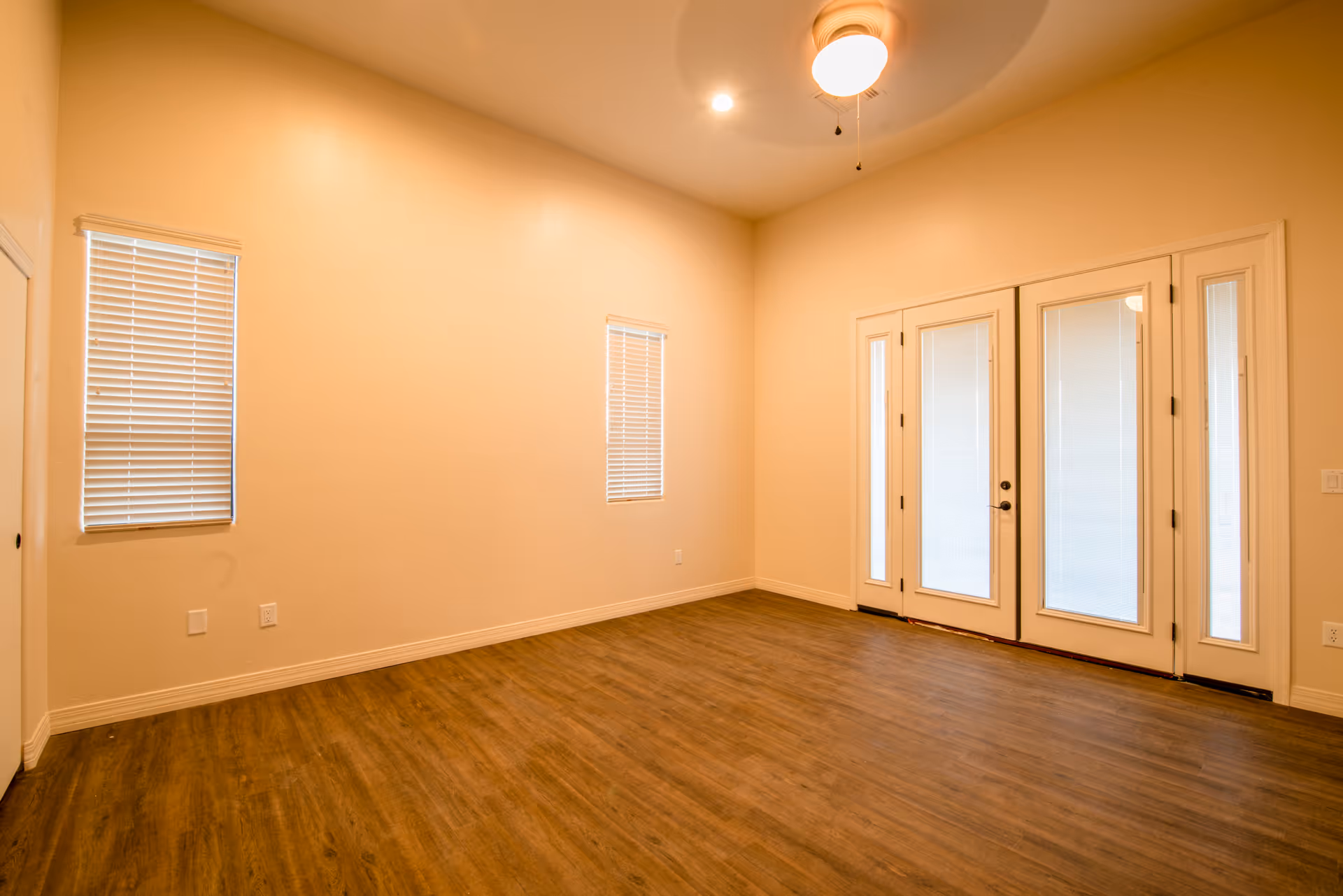 Empty room with wooden flooring, beige walls, two narrow windows with blinds, and a set of glass double doors with blinds. A ceiling fan with a light is mounted on the ceiling.