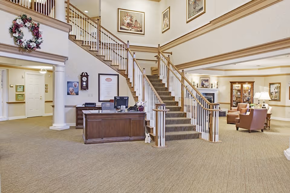 Interior view of a senior living facility lobby with a central wooden staircase carpeted in brown. A wooden reception desk with a computer and office supplies is positioned in front of the staircase. To the right, there is a seating area with brown leather chairs, a wooden cabinet with glass doors, a fireplace, and framed artwork on the walls. The space is well-lit with warm lighting and decorated with a floral wreath and paintings.