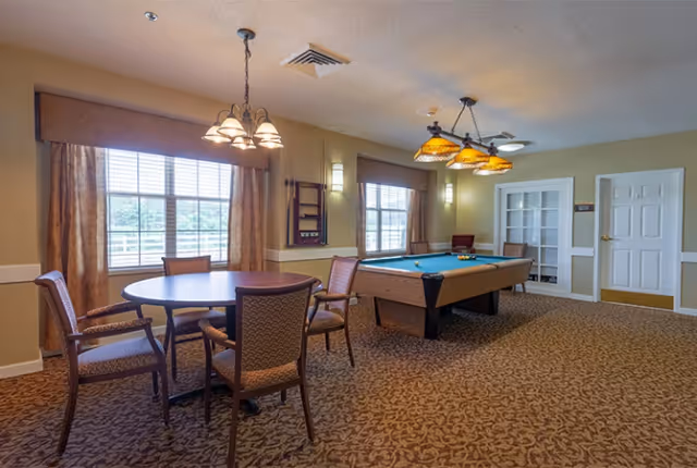 Carpeted common room with a pool table, a round table and chairs, large windows, and hanging light fixtures.
