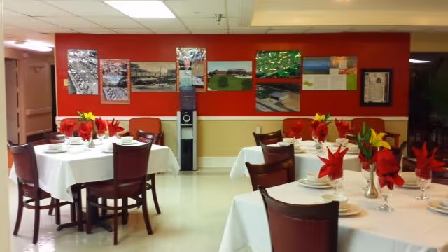 Dining room with tables covered in white tablecloths, set with plates, glasses, and red napkins folded decoratively. Chairs are arranged around the tables. The back wall is red with framed pictures and a water cooler in the center.