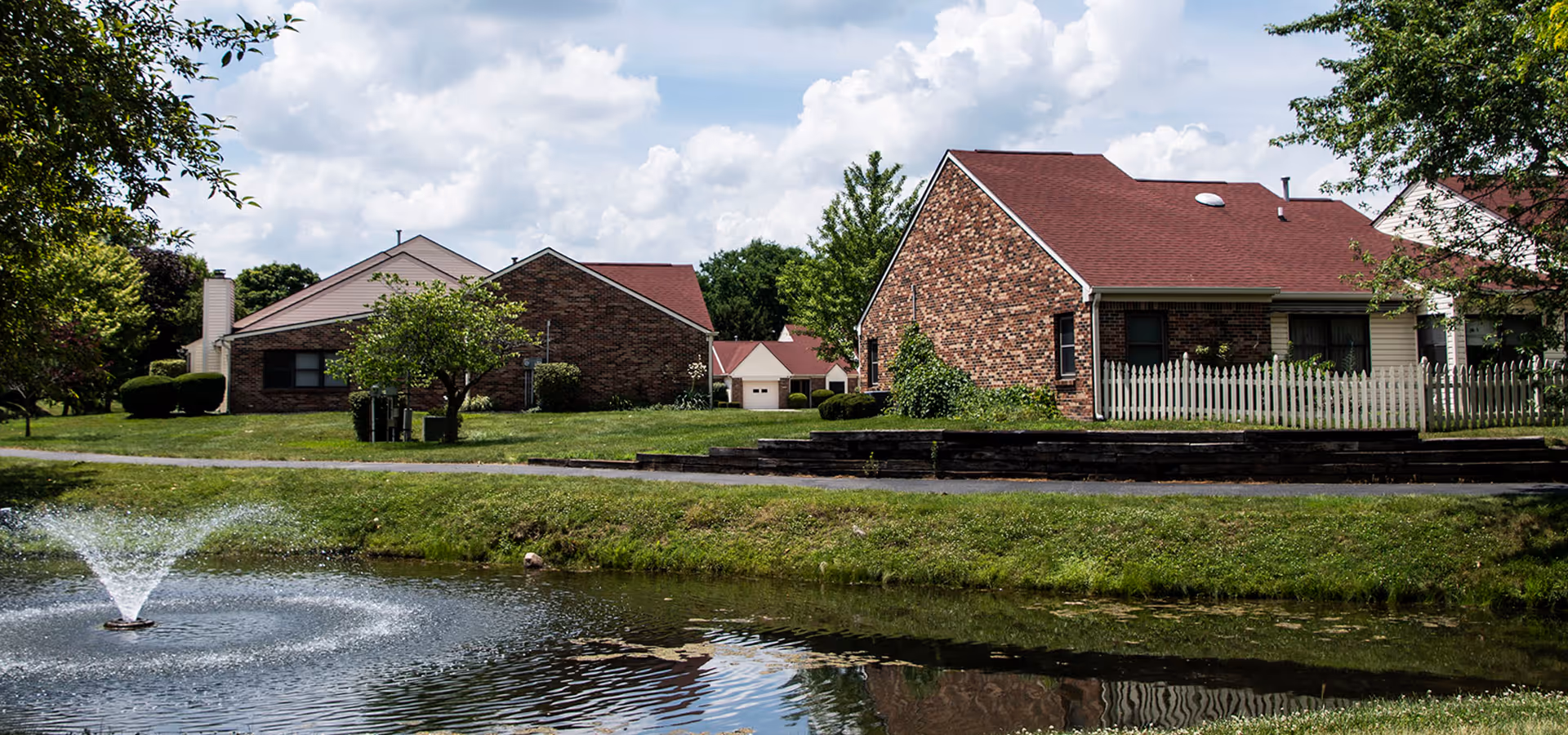 Brick and siding cottages with red roofs and a white picket fence beside a pond with a fountain under a partly cloudy sky.