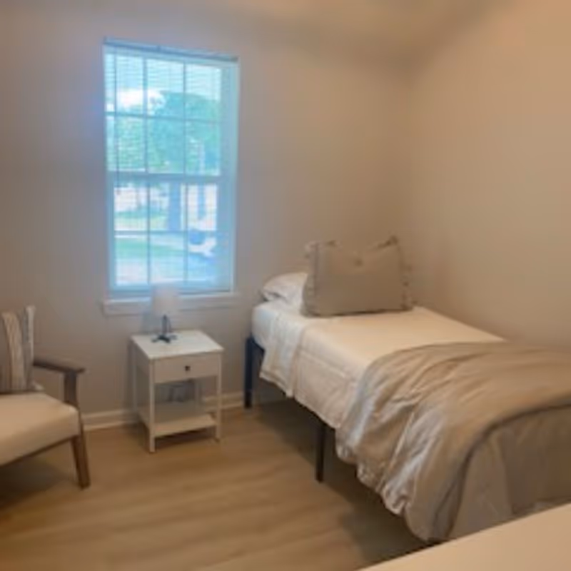 Small, minimally furnished bedroom with a single bed, nightstand and armchair beside a window.
