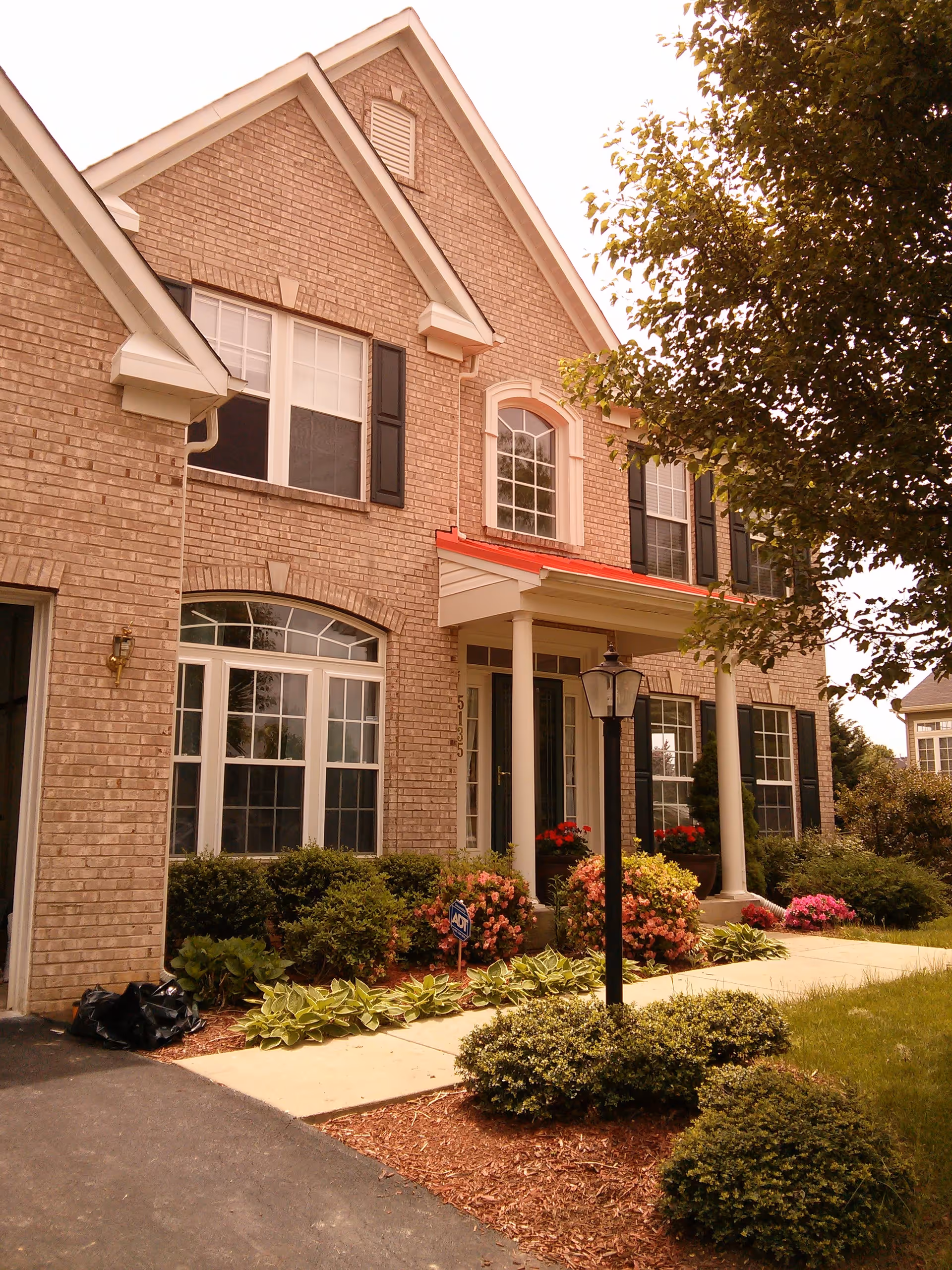 Front exterior view of a two-story brick house with white framed windows, black shutters, a red roof over the porch, and a black lamp post in the landscaped front yard with bushes and flowers.