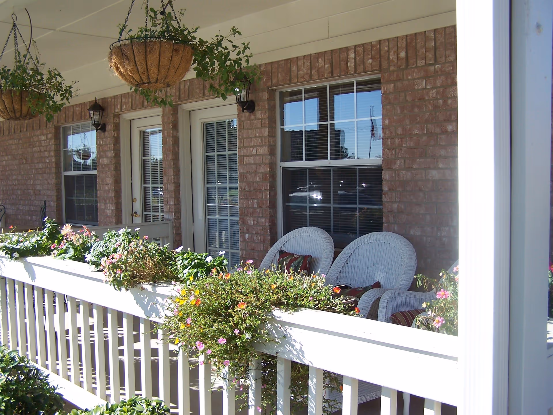 A porch area with white railing and flower boxes filled with colorful flowers. Two white wicker chairs with cushions are placed near a window and a door on a brick wall. Hanging baskets with green plants are suspended from the porch ceiling.