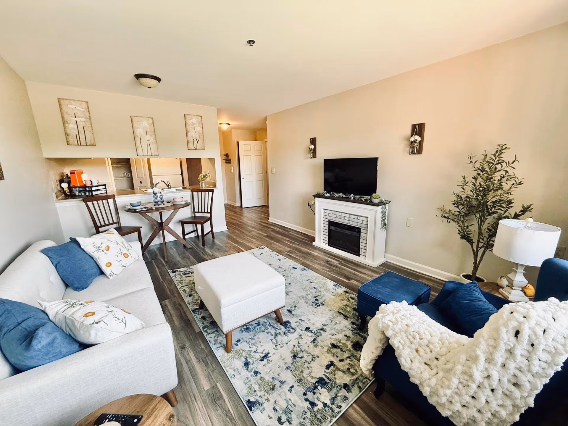 A cozy living room with a white sofa adorned with blue and floral pillows, a blue armchair with a white knitted throw, a white ottoman on a patterned rug, a small round dining table with two chairs, a TV mounted above a white electric fireplace, and a potted plant in the corner. The room has wooden flooring and beige walls with decorative wall hangings.