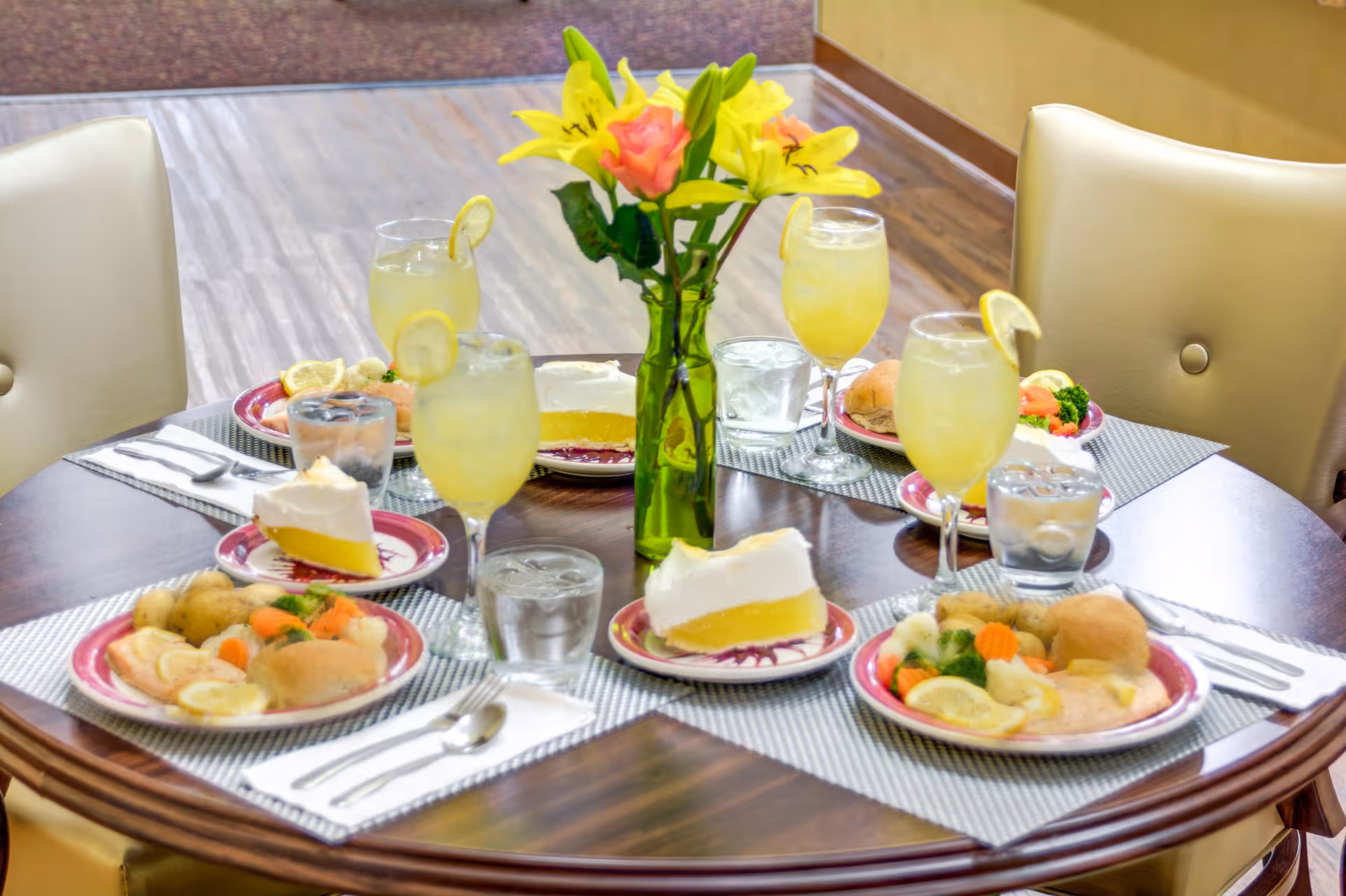 A round dining table set for four with plates of food including vegetables, rolls, and lemon meringue pie slices. Each place setting has a glass of water and a glass of lemonade with a lemon slice. A green vase with yellow lilies and a pink rose is in the center of the table. The table is in a room with wood flooring and beige cushioned chairs.