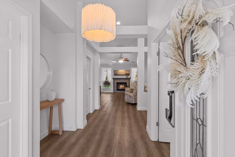 View down a hallway with wooden flooring leading to a living room area with a fireplace, armchairs, and windows with white curtains. The hallway has white walls, a modern ceiling light fixture, a small wooden console table with a decorative vase, and a door decorated with a white feather wreath.