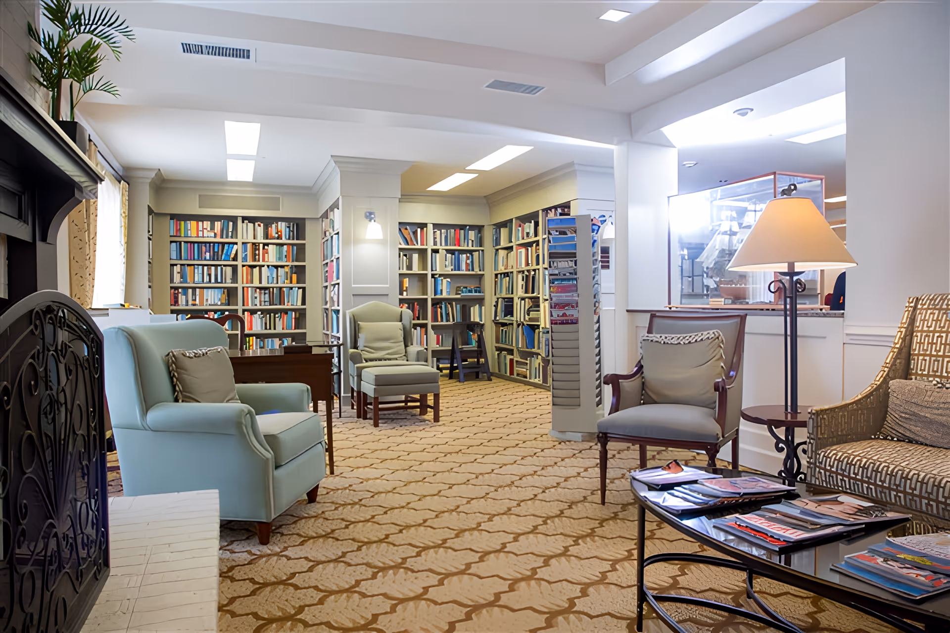 Comfortable common lounge with upholstered armchairs, bookshelves lining the back wall, a coffee table with magazines, and warm lamps.