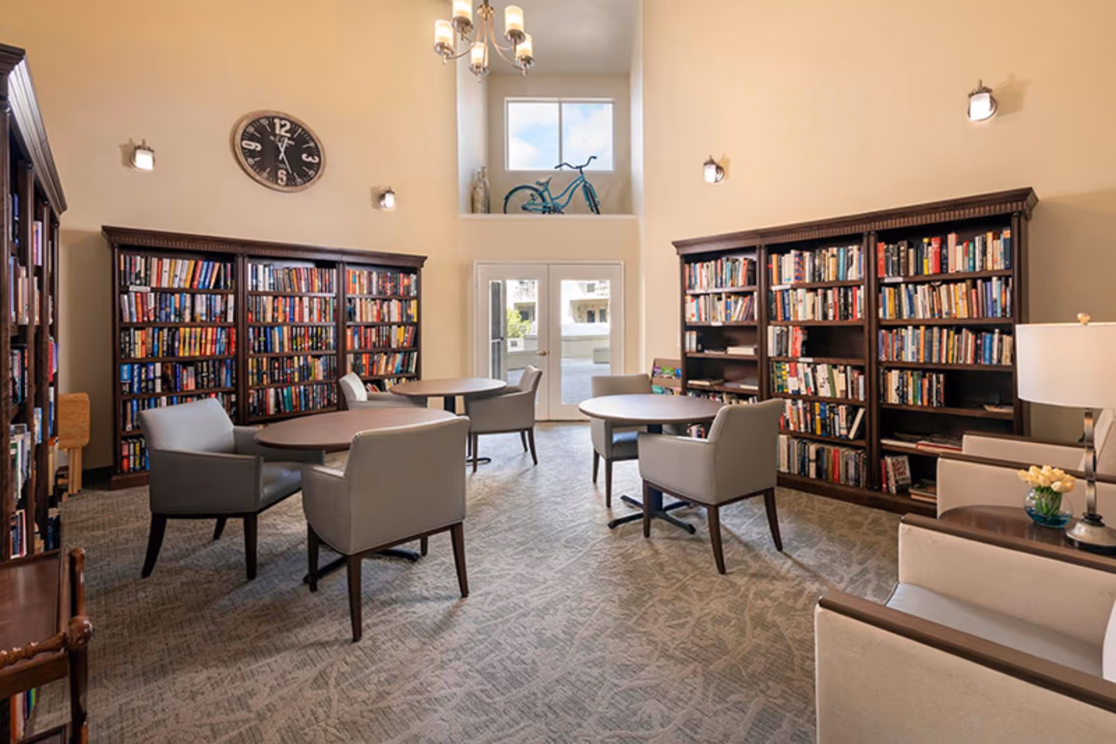 A cozy library room with multiple dark wood bookshelves filled with books, round tables surrounded by gray upholstered chairs, a large clock on the wall, a chandelier hanging from the ceiling, and a small decorative bicycle displayed on a ledge above double glass doors.