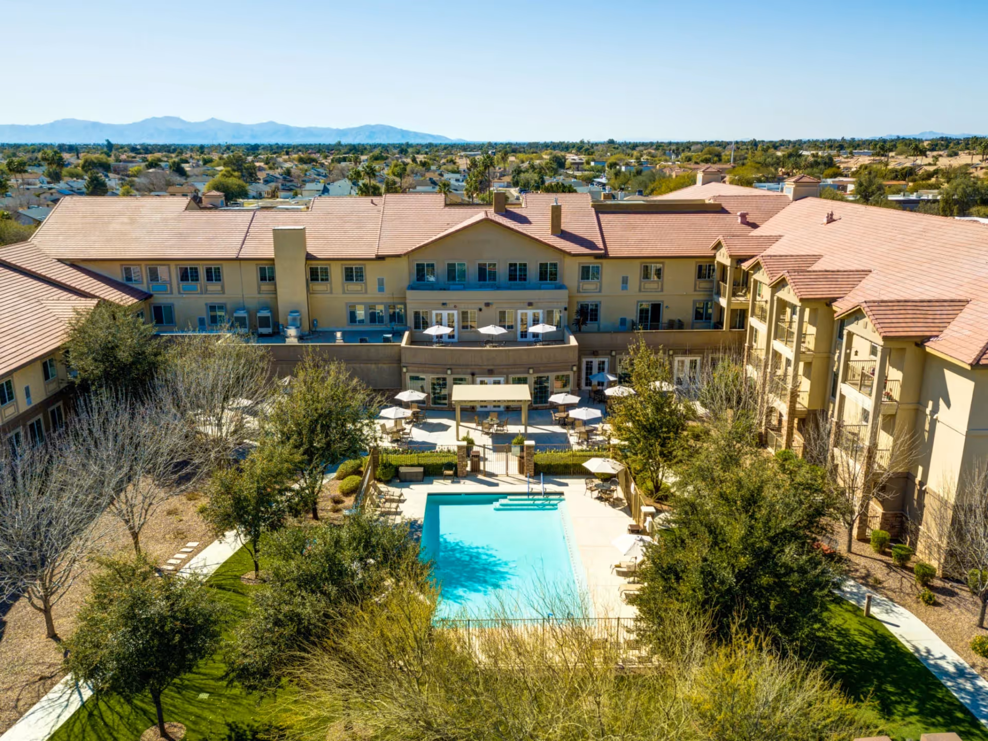 Aerial view of Palos Verdes Senior Living facility showing a large outdoor swimming pool surrounded by lounge chairs and umbrellas, with a multi-story building in the background and trees and greenery around the pool area.