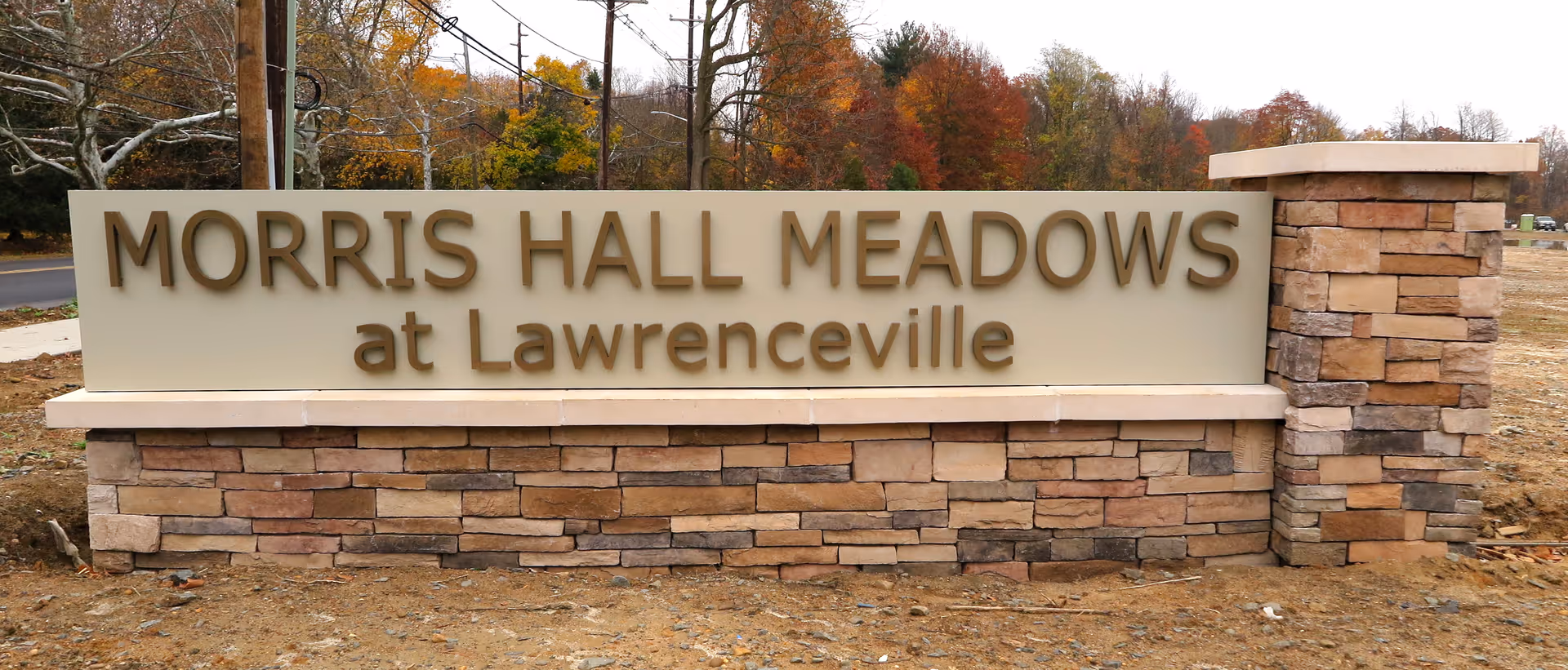 A large stone and concrete sign with the text 'MORRIS HALL MEADOWS at Lawrenceville' situated outdoors with trees and a road in the background.