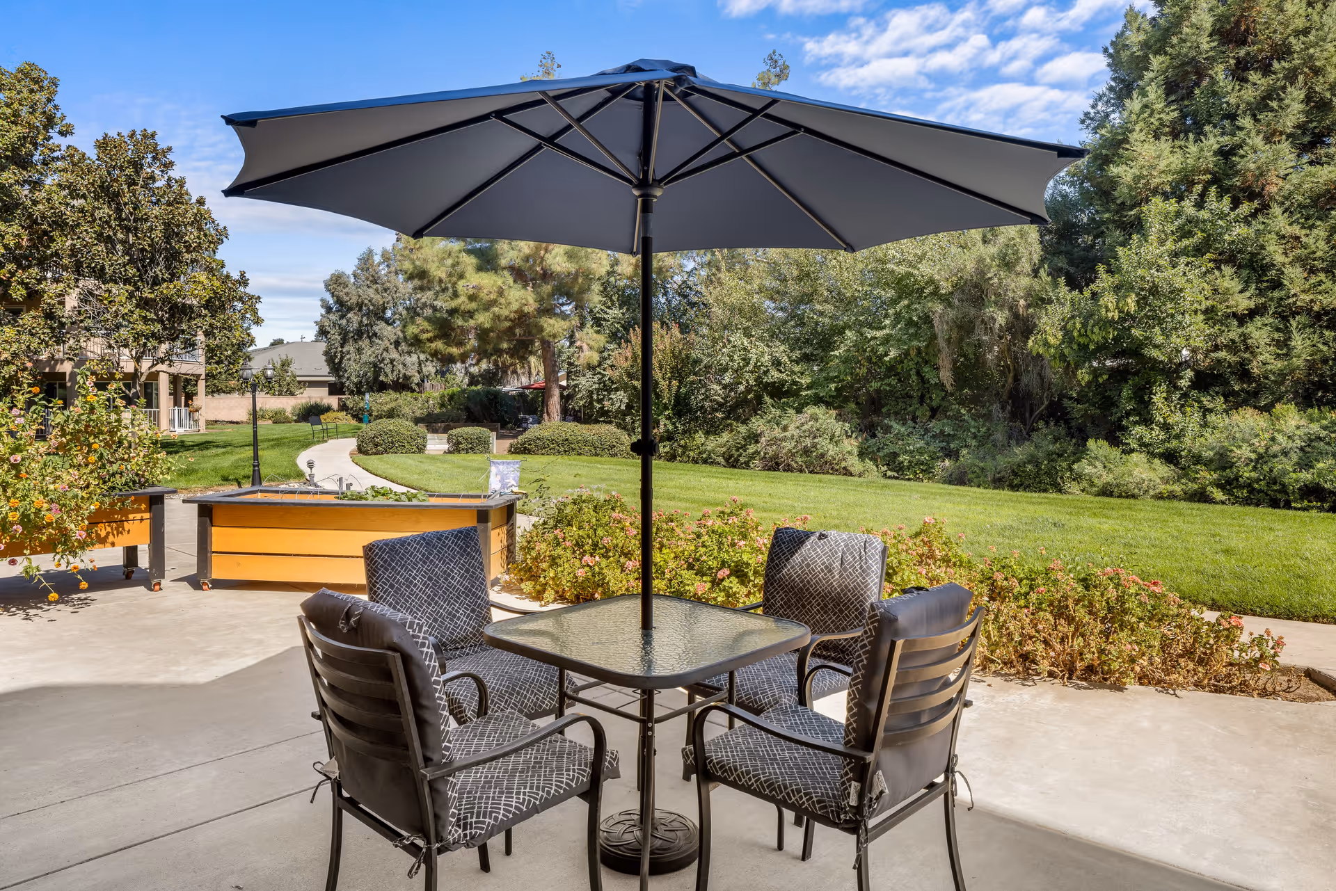 Outdoor patio area with a square glass table surrounded by four cushioned chairs under a large black umbrella. The patio overlooks a green lawn, bushes, and trees under a partly cloudy blue sky.
