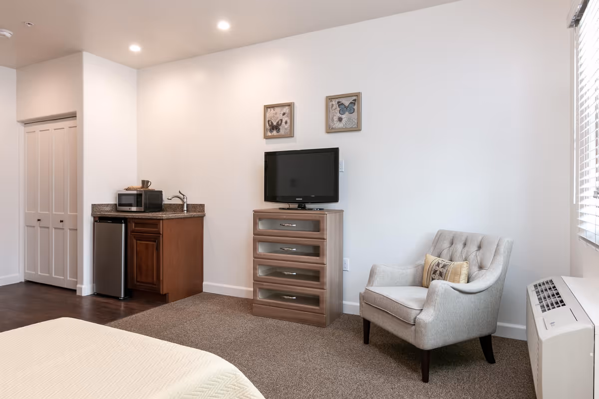 A cozy studio bedroom with a small kitchenette and mini fridge, a TV on a dresser, an upholstered armchair, and a partially visible bed.
