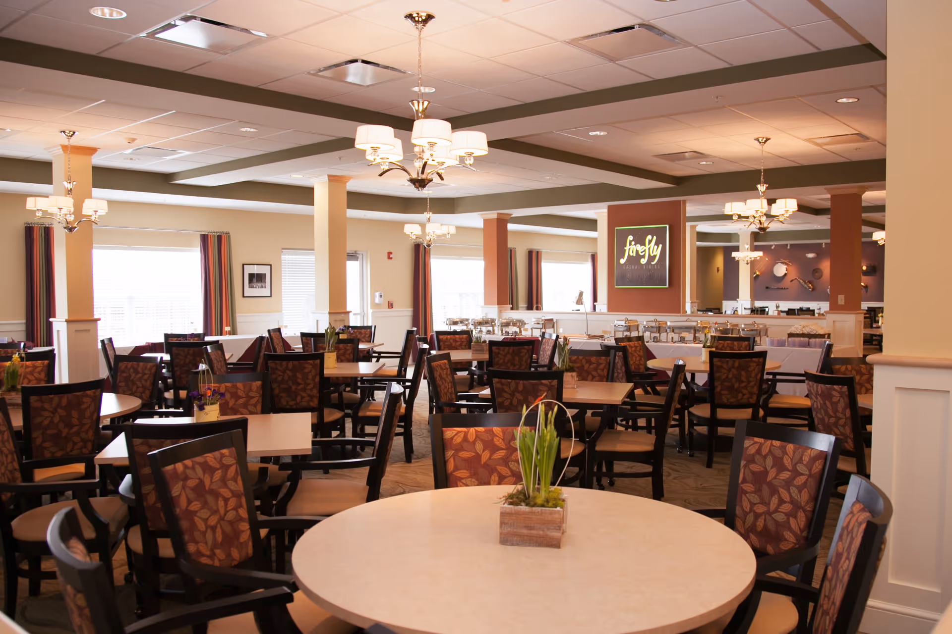A spacious dining room with multiple round and square tables surrounded by chairs with patterned upholstery. The room is well-lit with several chandeliers hanging from the ceiling. There are windows with curtains along the walls, and a sign on the far wall reads 'firefly'. The overall decor is warm and inviting.