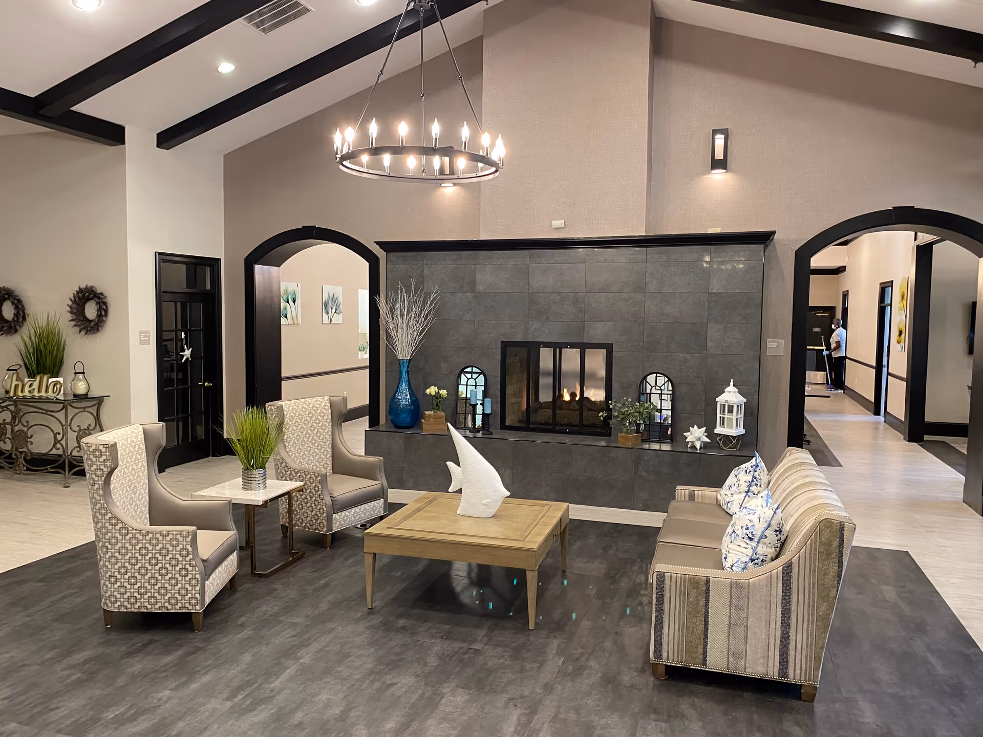 Communal lounge with armchairs and a sofa arranged around a coffee table in front of a tiled fireplace under a chandelier.